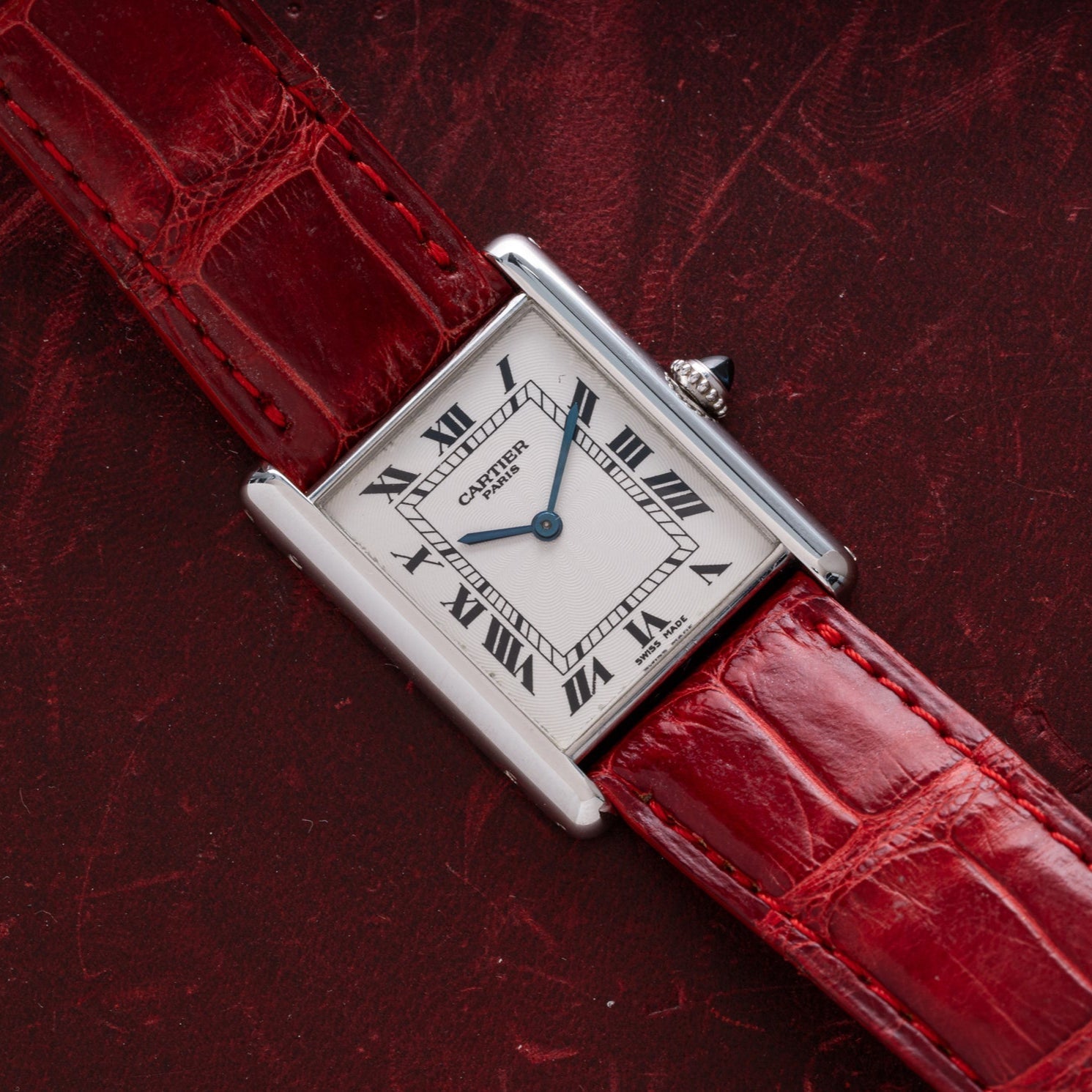 A Cartier Tank Louis watch with a red leather strap displayed on a dark red textured surface.