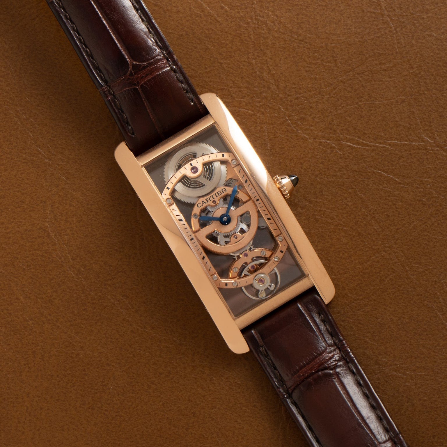 The Cartier Tank Cintreé Skeleton is a rectangular gold watch with a skeleton dial and brown leather strap, displayed on a brown leather surface.