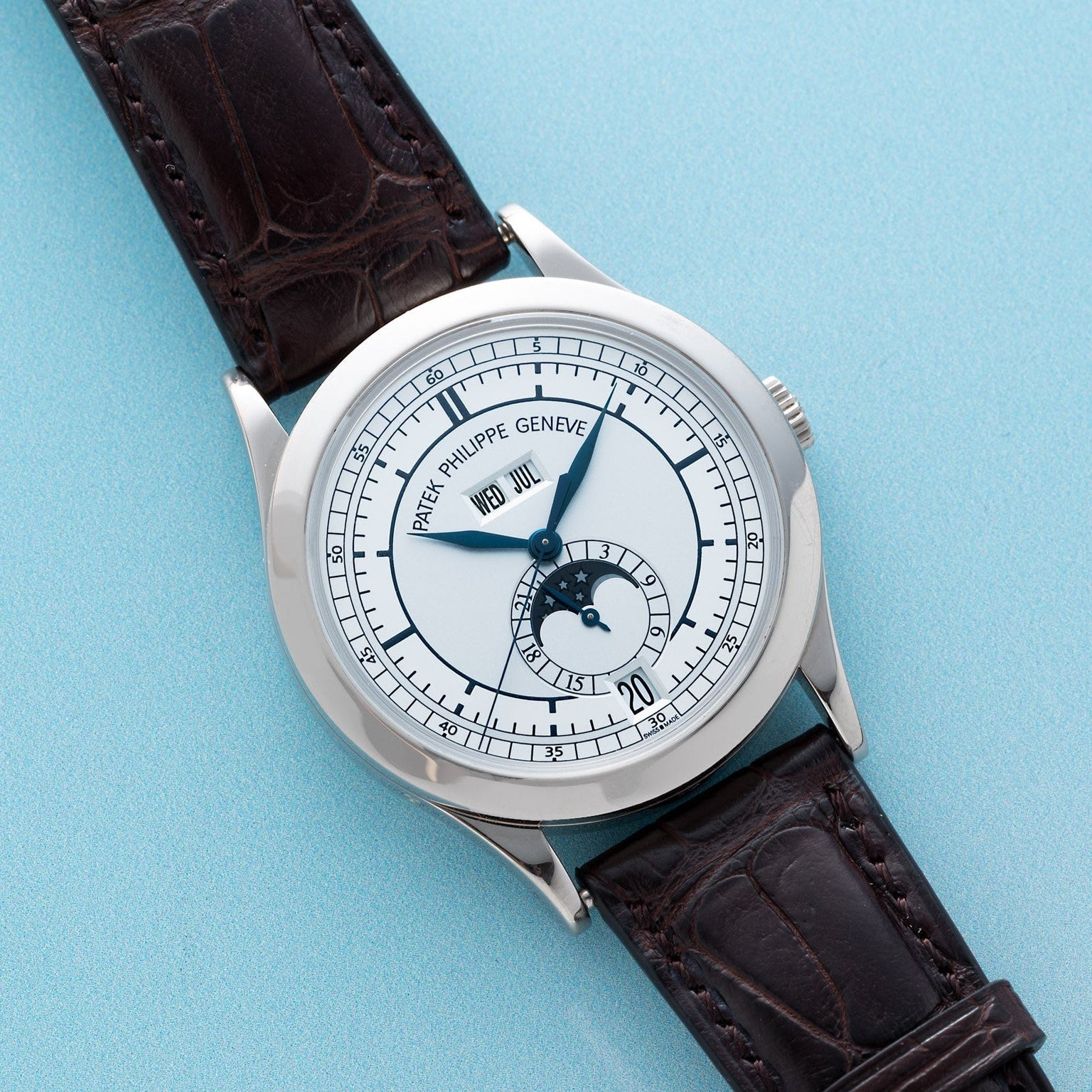 Patek Philippe Annual Calendar watch with white face, moonphase indicator, and dark brown leather strap displayed on a light blue background.