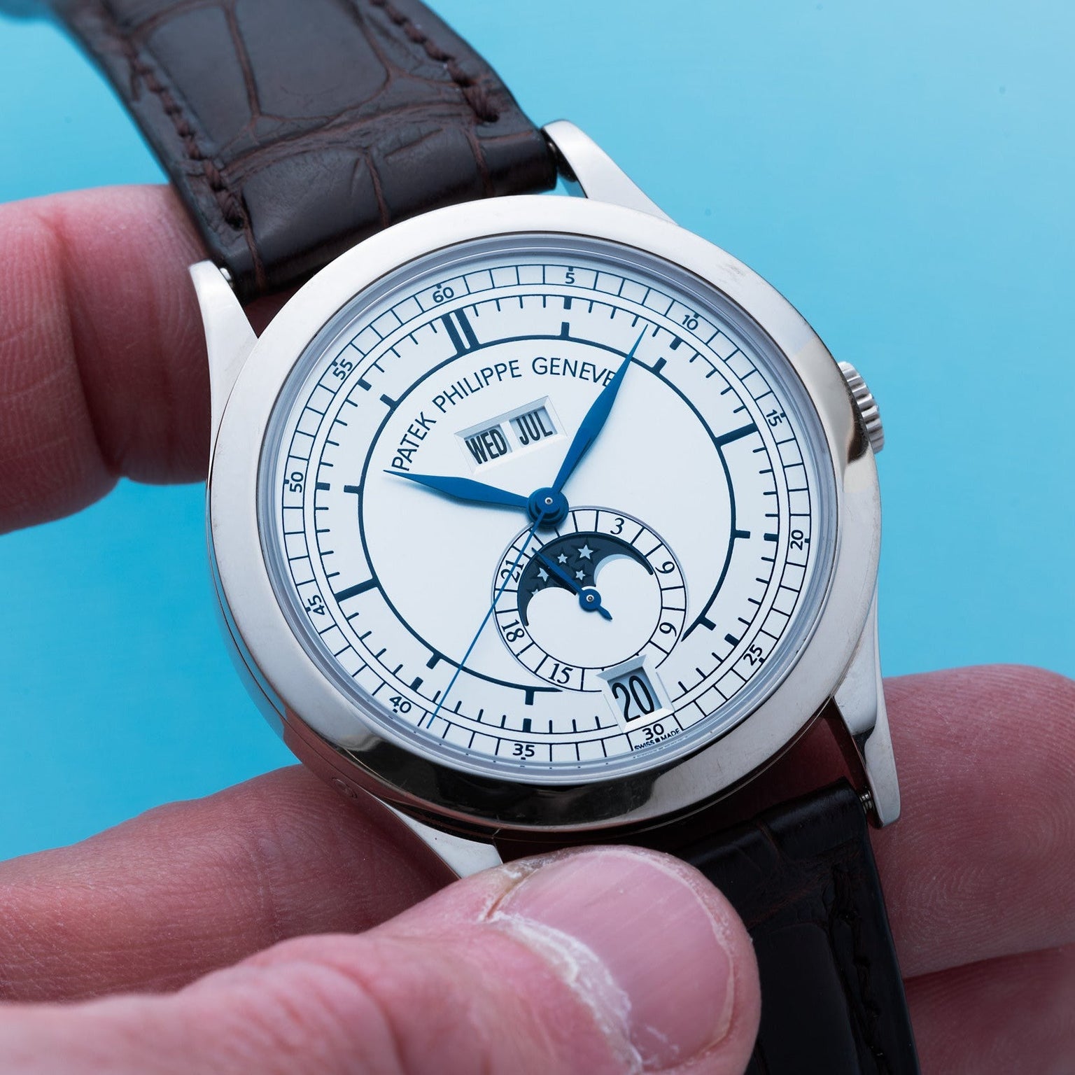 A hand holds a Patek Philippe Annual Calendar wristwatch featuring a moon phase and date display on a blue background.