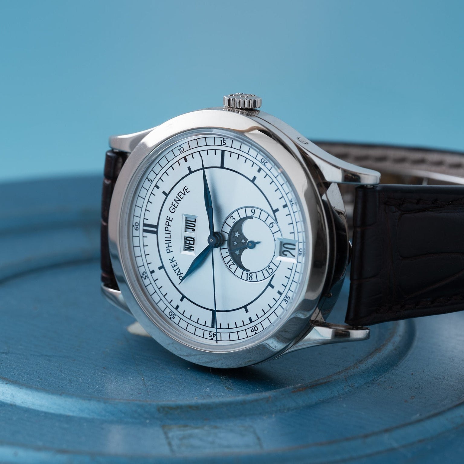 The Patek Philippe Annual Calendar luxury wristwatch features a white dial, black leather strap, and calendar display, shown on a blue surface.