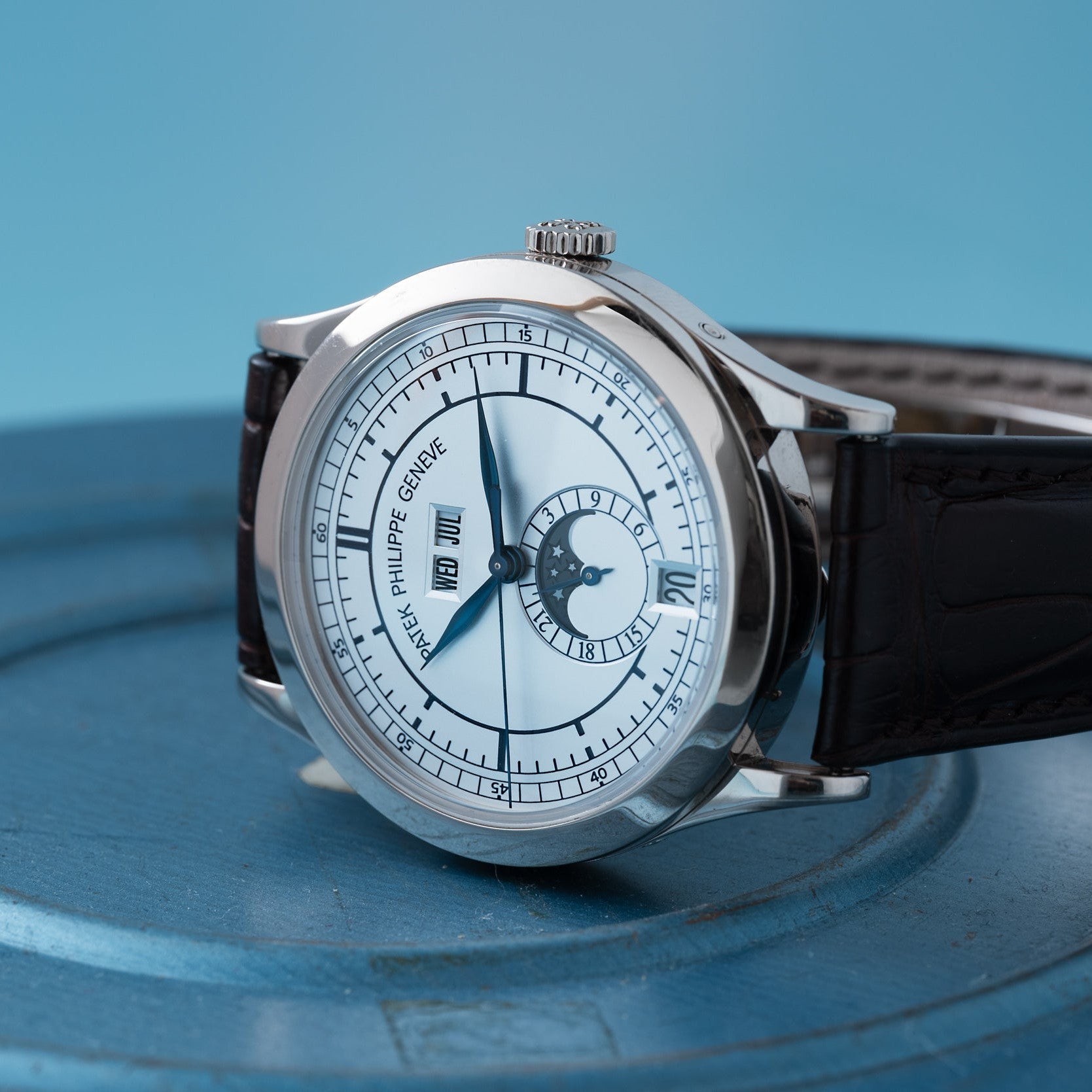 The Patek Philippe Annual Calendar luxury wristwatch features a white dial, black leather strap, and calendar display, shown on a blue surface.