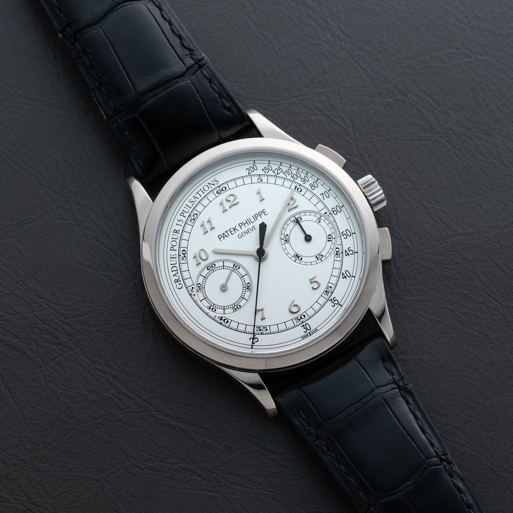 A Patek Philippe Chronograph wristwatch with a white dial and black leather strap displayed on a dark surface.