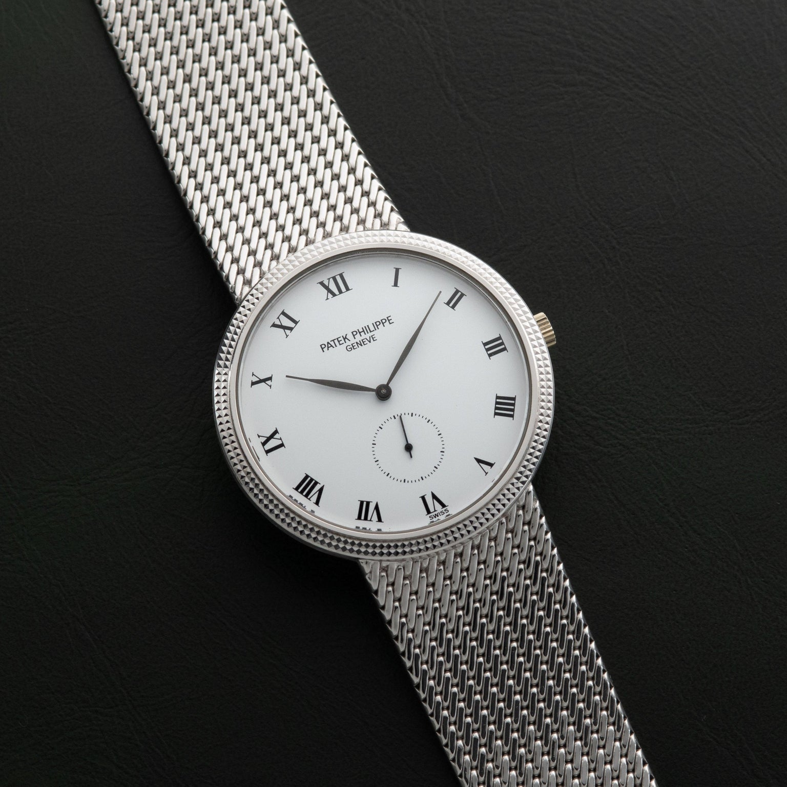 The Patek Philippe Calatrava is a silver mesh wristwatch featuring a white face with Roman numerals on a black background.
