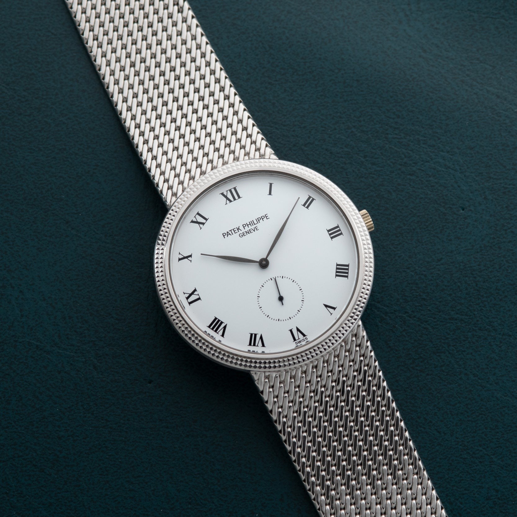 Patek Philippe Calatrava silver mesh watch with white face, Roman numerals, and classic design displayed on a dark green textured surface.