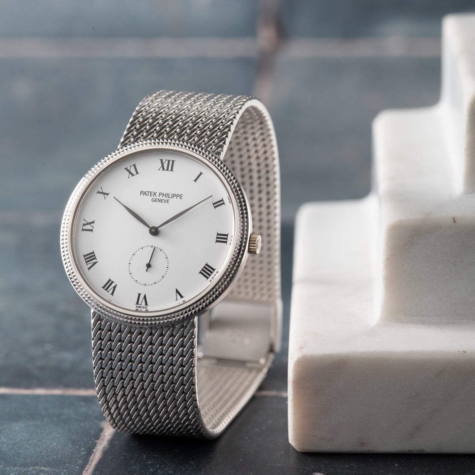 A silver Patek Philippe Calatrava watch with Roman numerals rests on a marble surface against a tiled background.