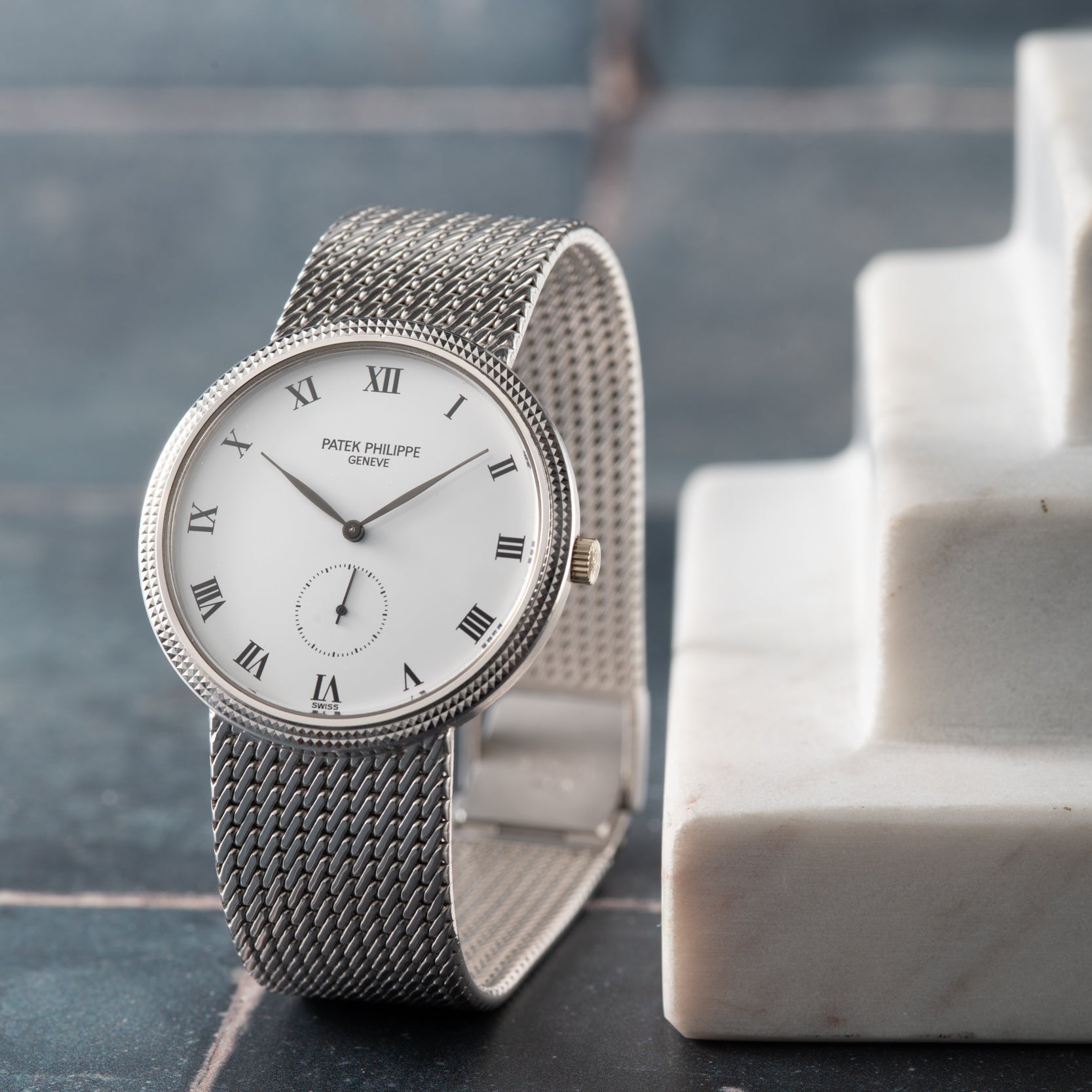 A silver Patek Philippe Calatrava watch with Roman numerals rests on a marble surface against a tiled background.