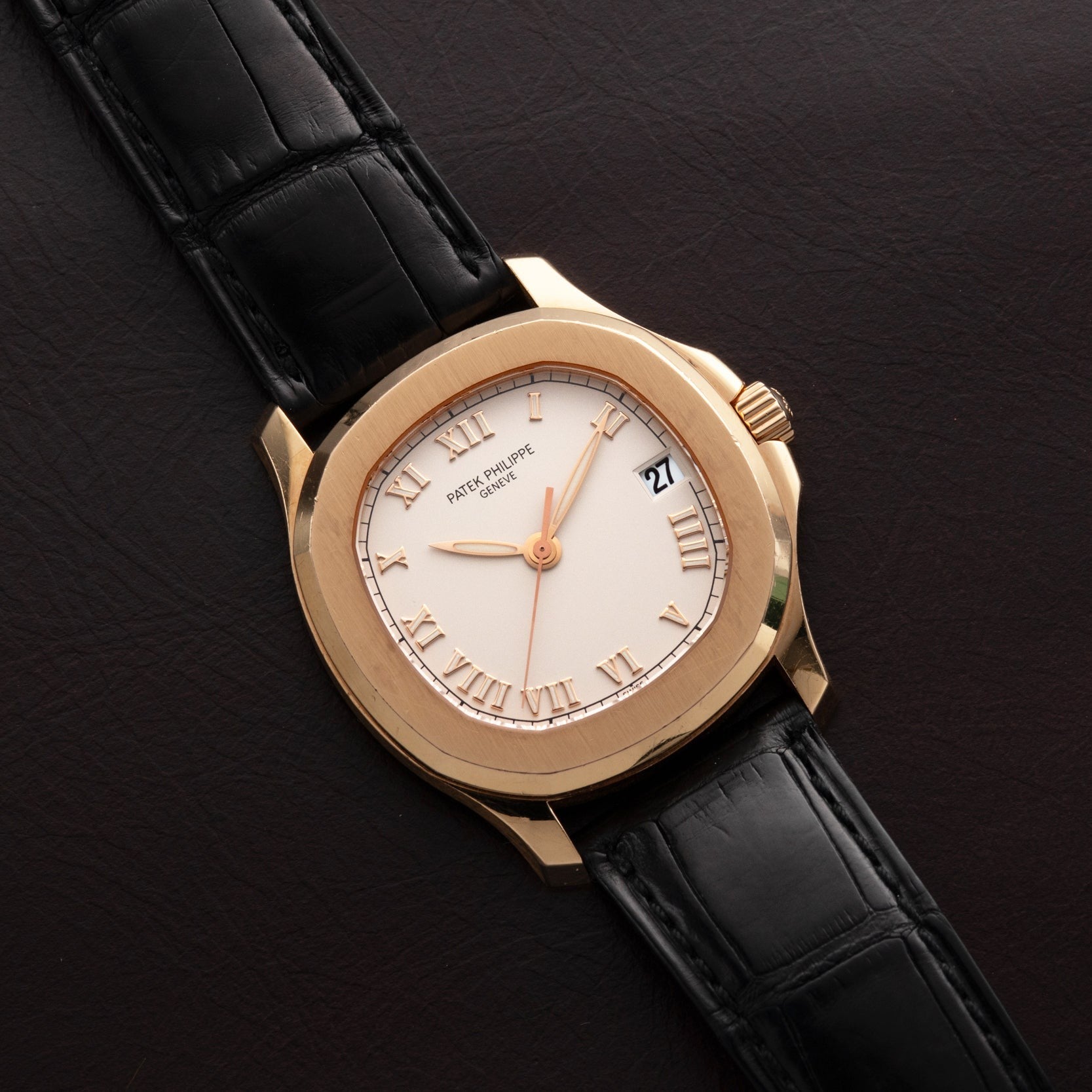 The Patek Philippe Aquanaut gold wristwatch with black leather strap rests on a dark brown leather surface, displaying Roman numerals and date.