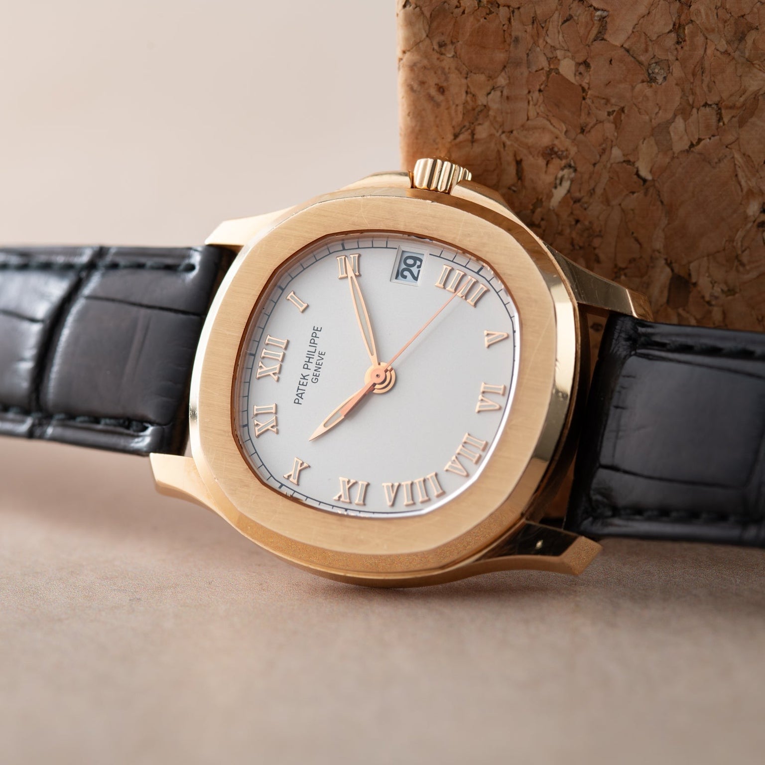 A Patek Philippe Aquanaut gold wristwatch with a white dial, Roman numerals, and a black leather strap rests on a cork surface.