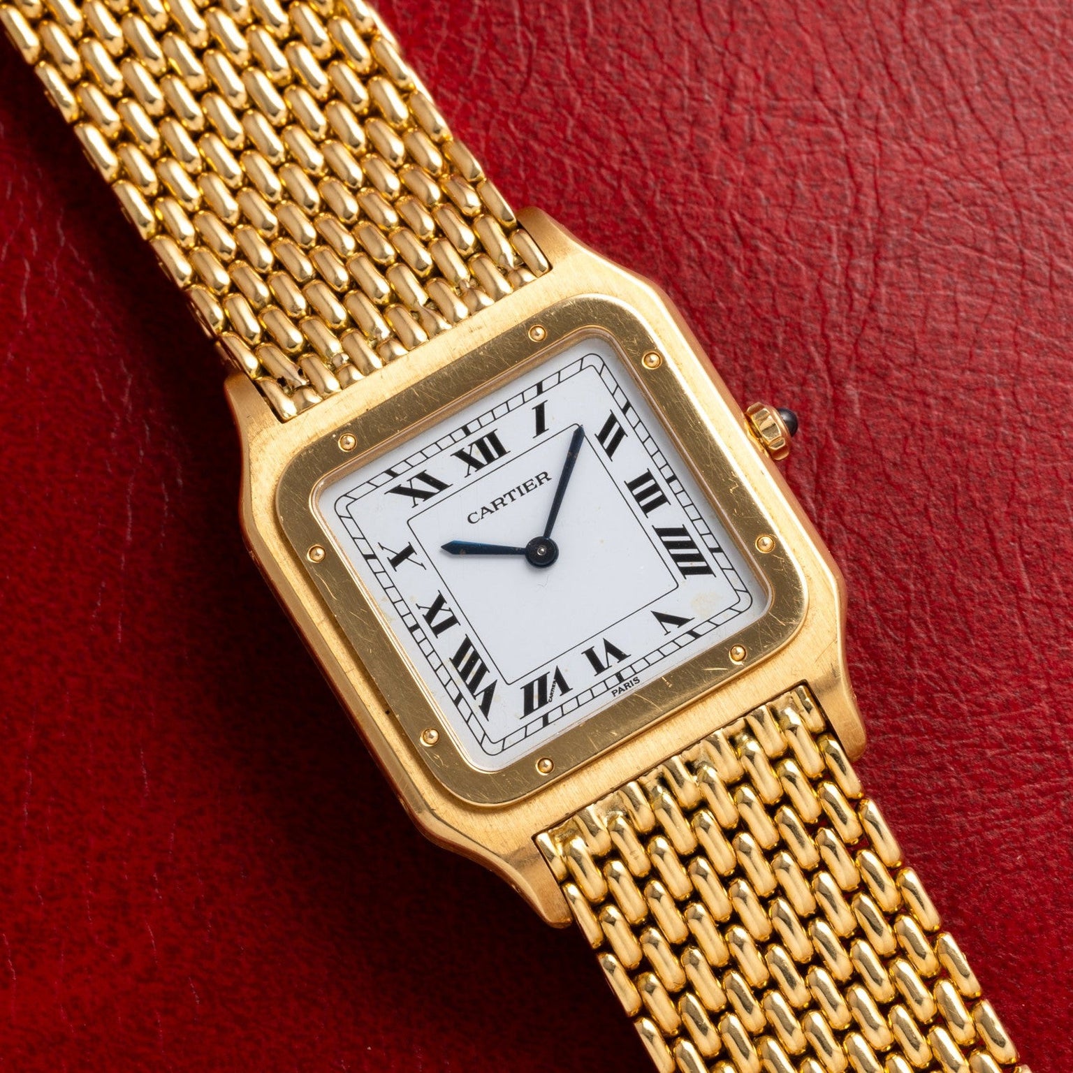 The Cartier Santos Dumont is a gold watch featuring a square face with Roman numerals on a red leather strap.