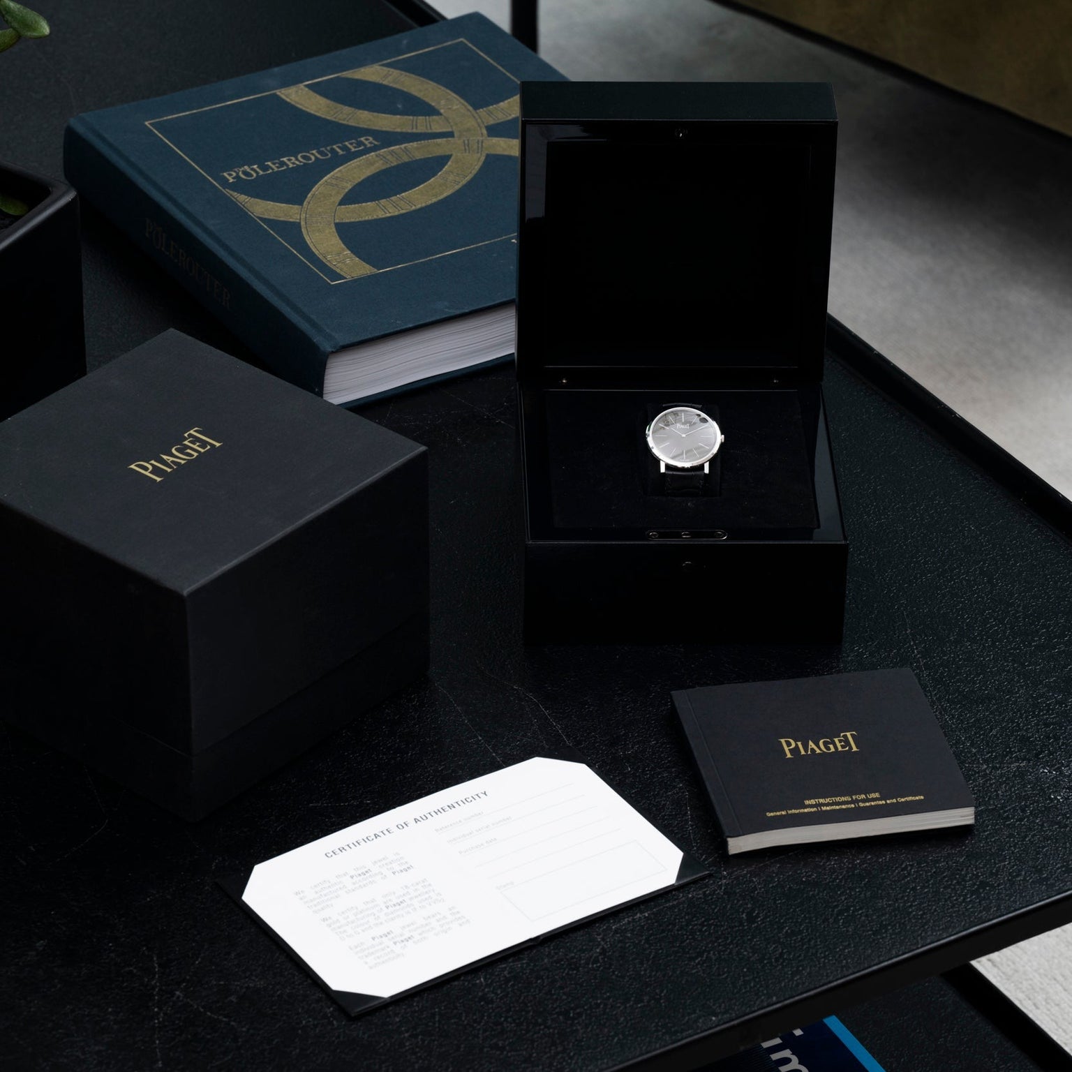 A Piaget Altiplano luxury watch in a black box sits next to a certificate, booklet, and book on a dark table.