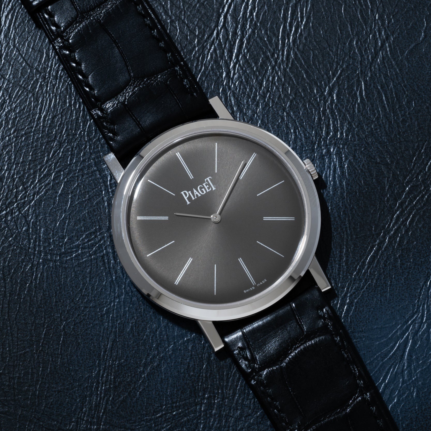 The Piaget Altiplano, a silver wristwatch with a black leather strap, is displayed on a textured dark surface.