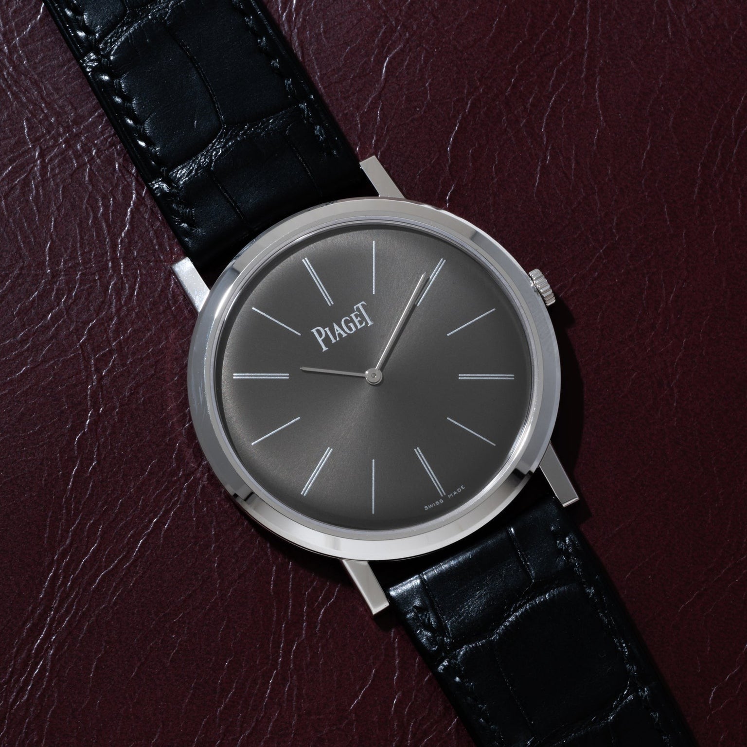 A Piaget Altiplano wristwatch with a black leather strap and minimalist black dial rests on a dark maroon leather surface.