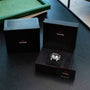 A TUDOR Black Bay Chronograph wristwatch rests in an open black box with a warranty card, alongside the outer box on a dark table.