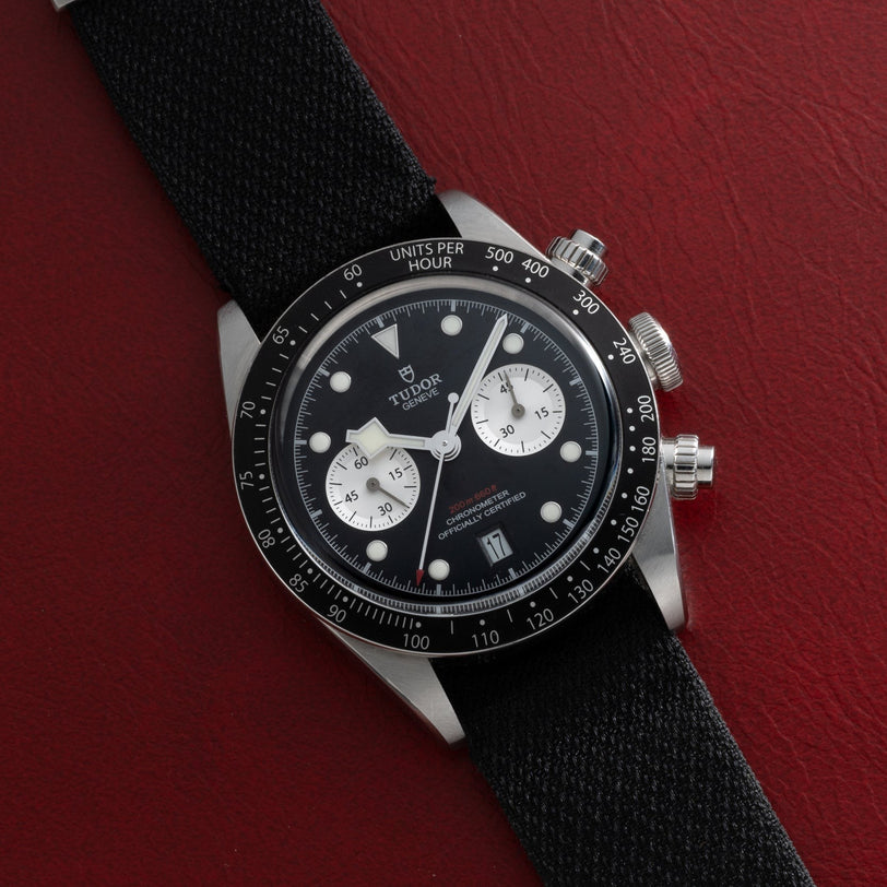 A TUDOR Black Bay Chronograph watch with a black strap displayed on a red textured surface.