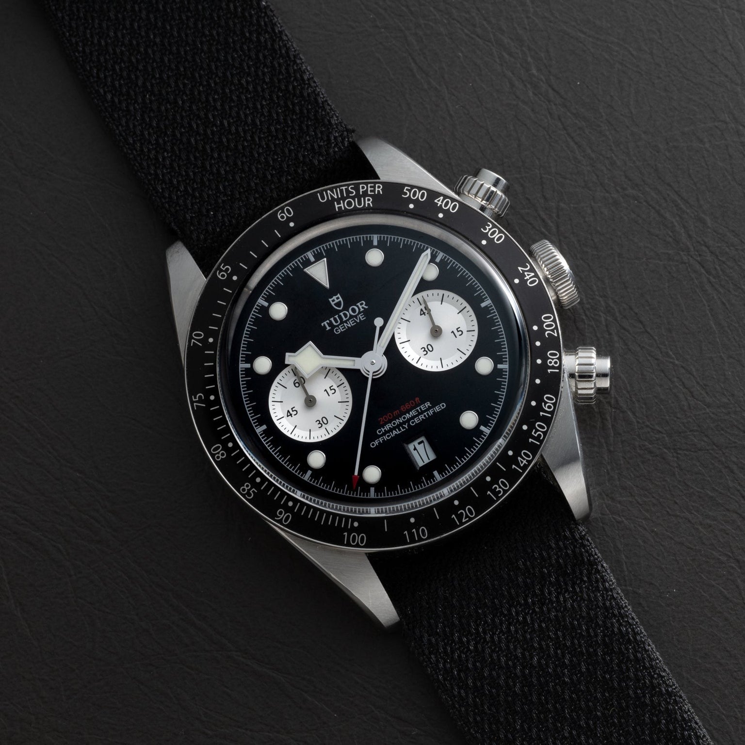 A TUDOR Black Bay Chronograph watch featuring a black dial, white subdials, and a black fabric strap displayed on a dark surface.