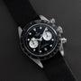 A TUDOR Black Bay Chronograph watch featuring a black dial, white subdials, and a black fabric strap displayed on a dark surface.