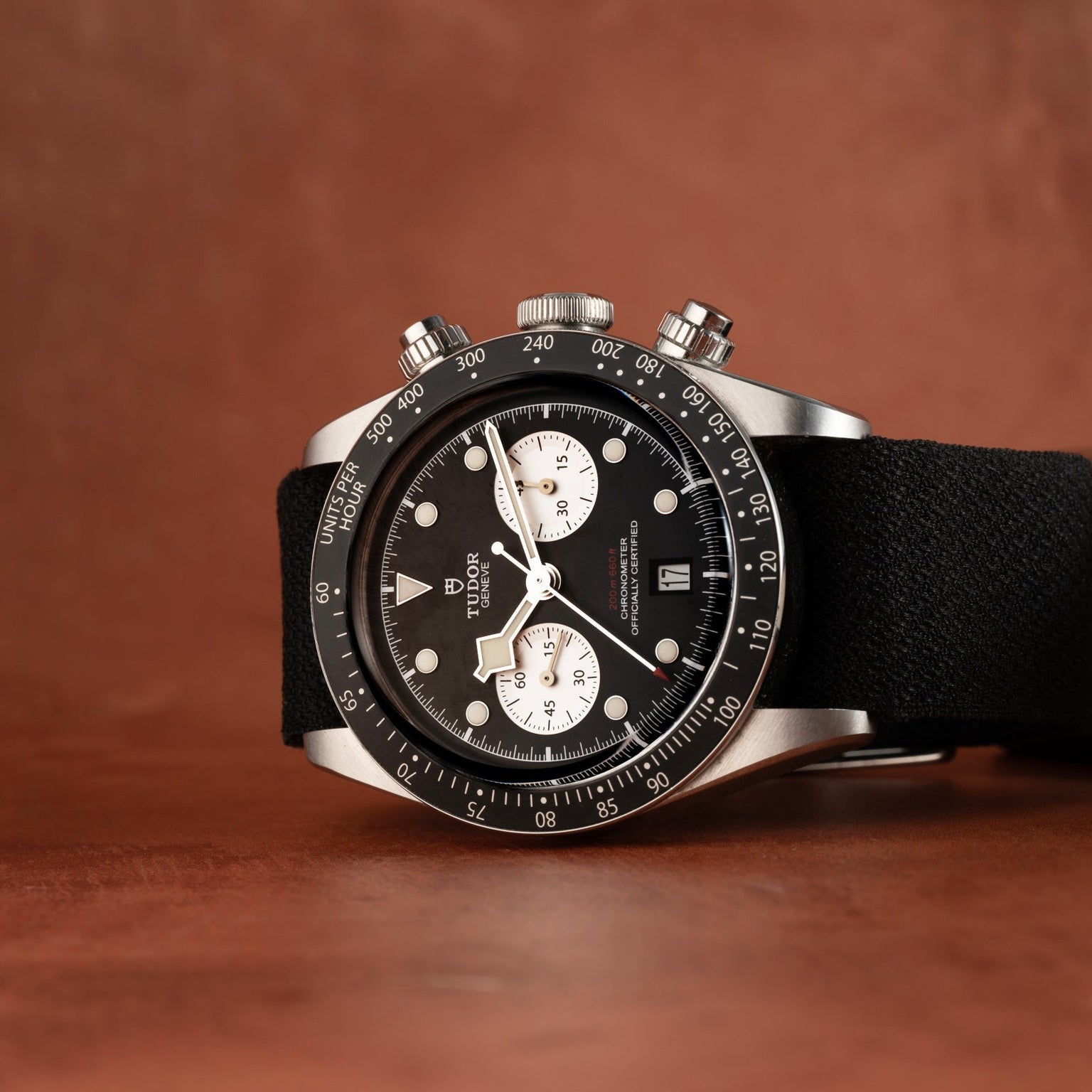 A TUDOR Black Bay Chronograph watch in black and silver with a fabric strap, placed on a brown surface.