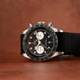 A TUDOR Black Bay Chronograph watch in black and silver with a fabric strap, placed on a brown surface.