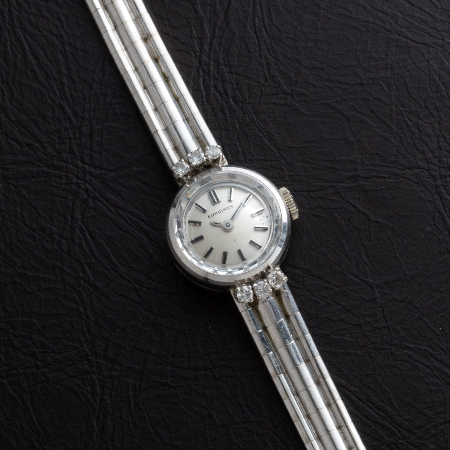 The Longines Diamond Cocktail Watch features a round face and striped silver band, elegantly showcased against a black textured background.