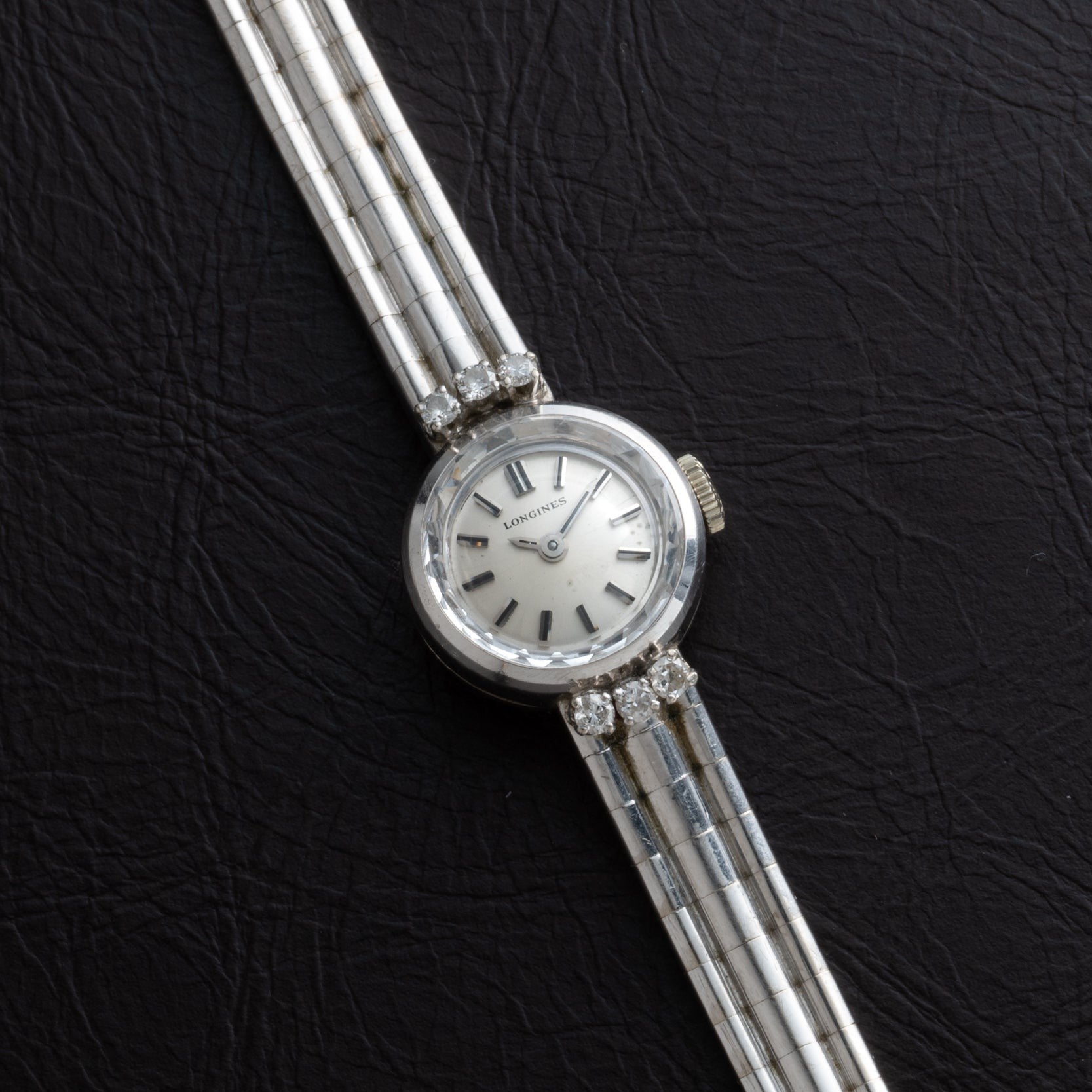 The Longines Diamond Cocktail Watch features a round face and striped silver band, elegantly showcased against a black textured background.