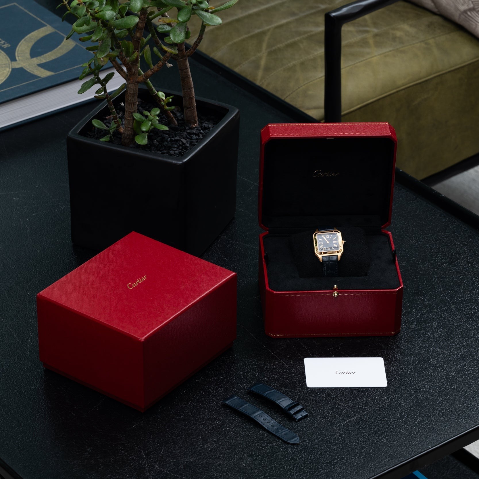 A Cartier Santos Dumont watch in its red box with packaging and card, displayed on a black table next to a plant.