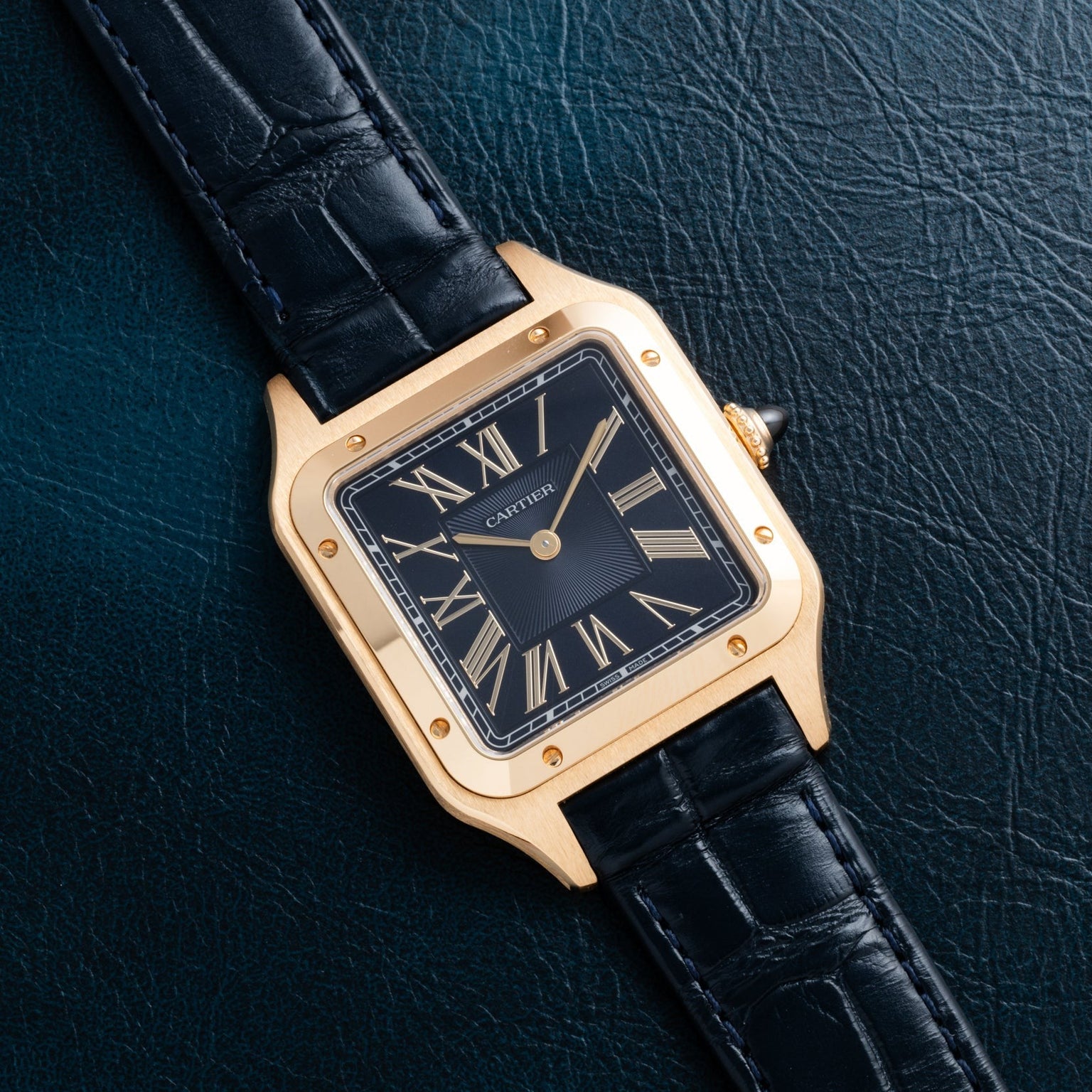 Cartier Santos Dumont gold watch with black leather strap, black dial, and Roman numerals displayed on a dark textured surface.