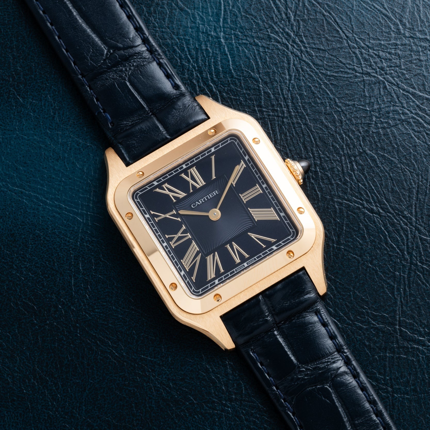 Cartier Santos Dumont gold watch with black leather strap, black dial, and Roman numerals displayed on a dark textured surface.