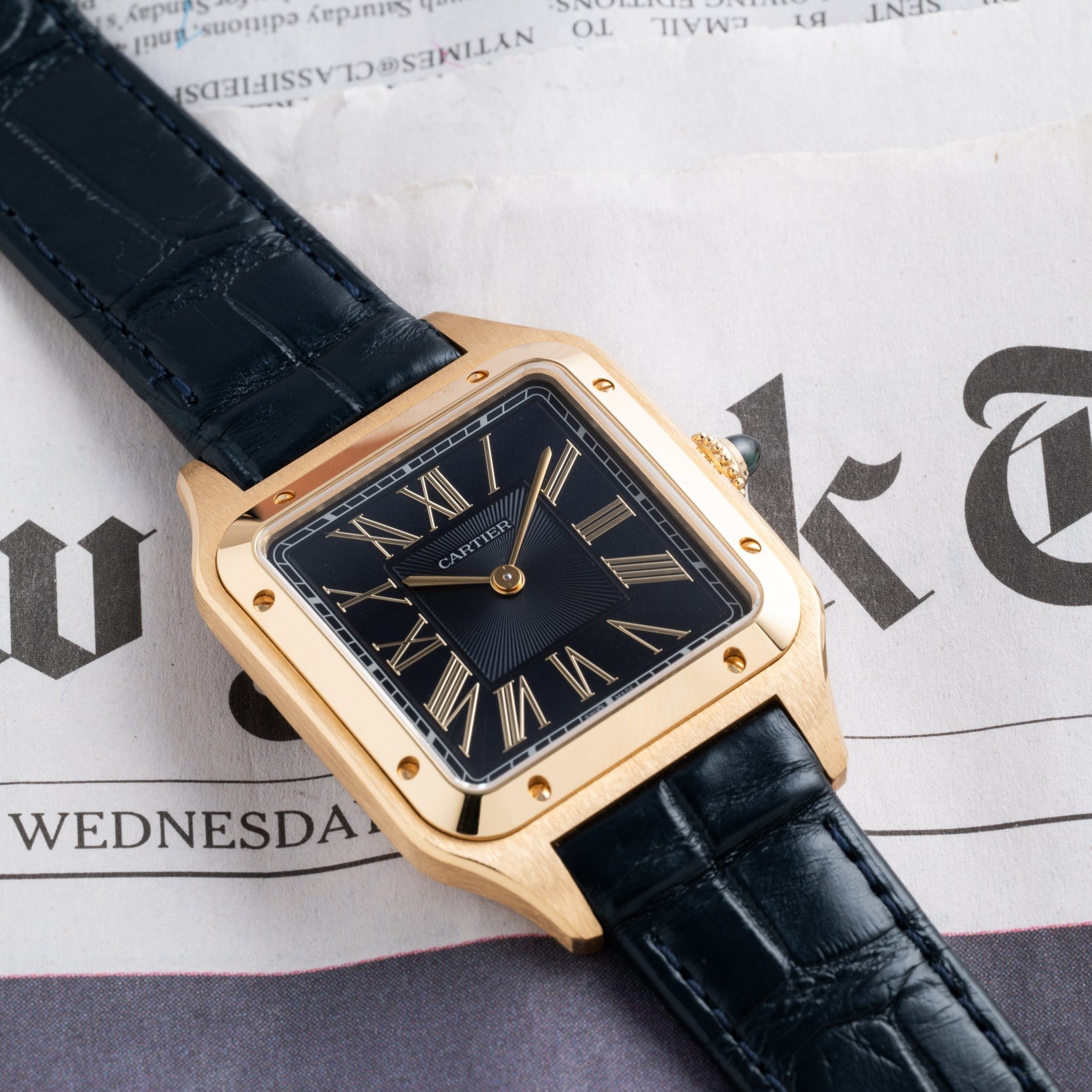 The Cartier Santos Dumont gold wristwatch with a black leather band rests on a newspaper displaying part of the word Wednesday.