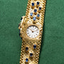 Patek Philippe Yellow Gold Integrated Dress Watch with blue gemstone accents on a green fabric background. The watch features a small, round face.