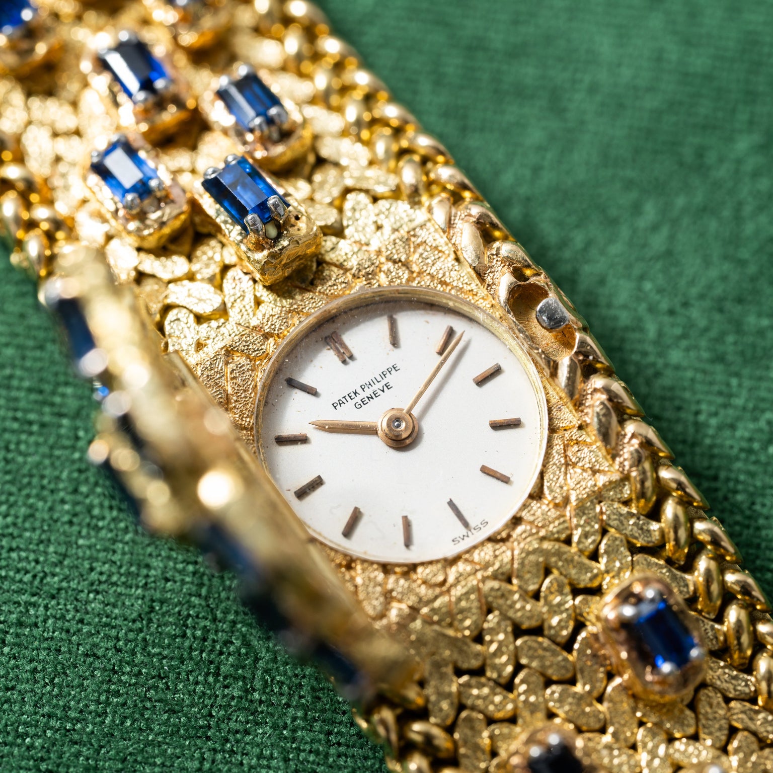 A Patek Philippe Yellow Gold Integrated Dress Watch displayed on a textured green fabric background with blue gemstones.