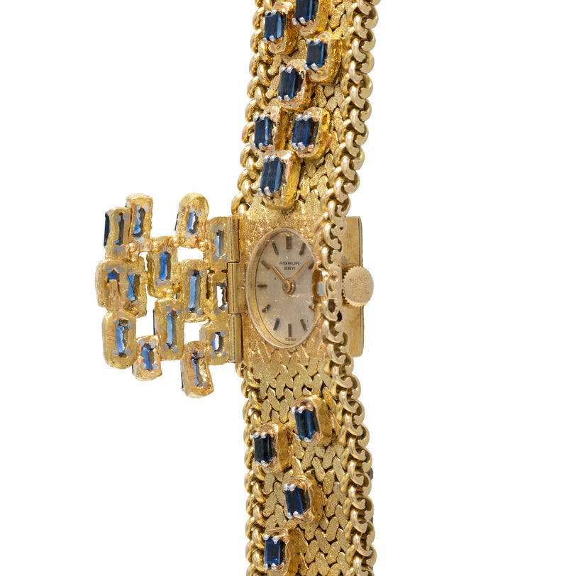 The Patek Philippe Yellow Gold Integrated Dress Watch features a textured band and blue gemstones set in a decorative bow design.