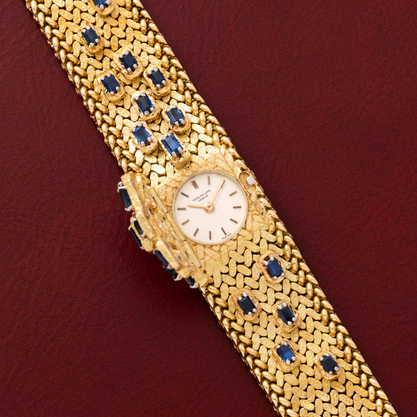 Patek Philippe Yellow Gold Integrated Dress Watch, featuring a textured band and blue gemstones, displayed on a dark red leather surface.