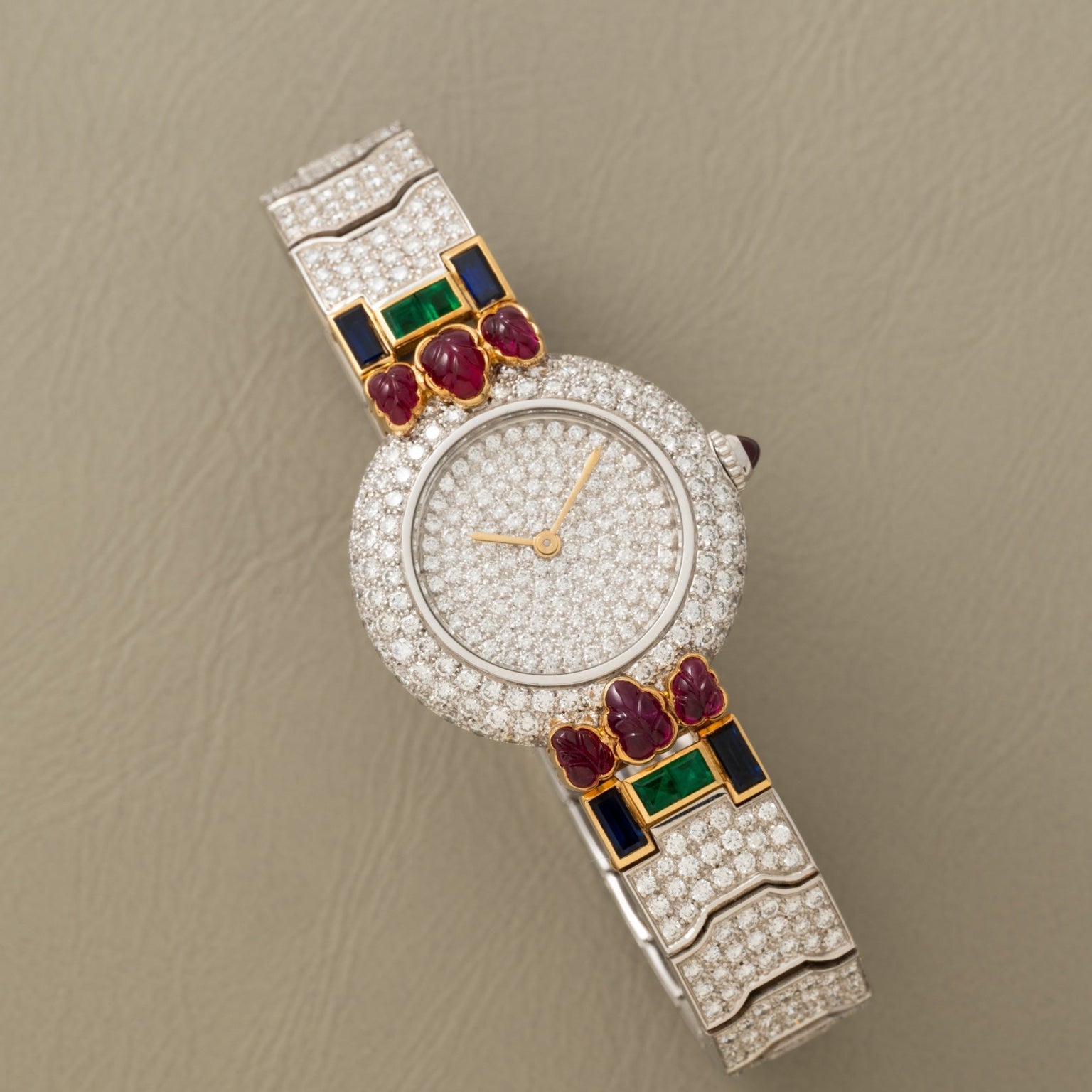 The Cartier Colisee Tutti Frutti Diamond watch features gold, ruby, emerald, and sapphire accents set against a beige background.