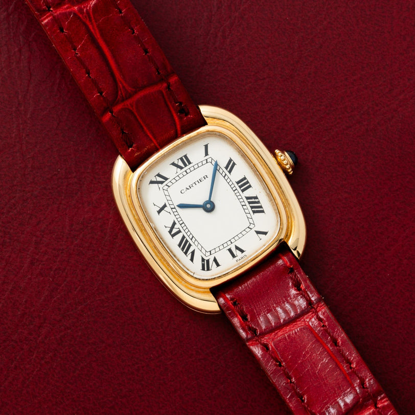 A Cartier Gondole Ladies gold watch with a red leather strap rests on a matching red leather surface.