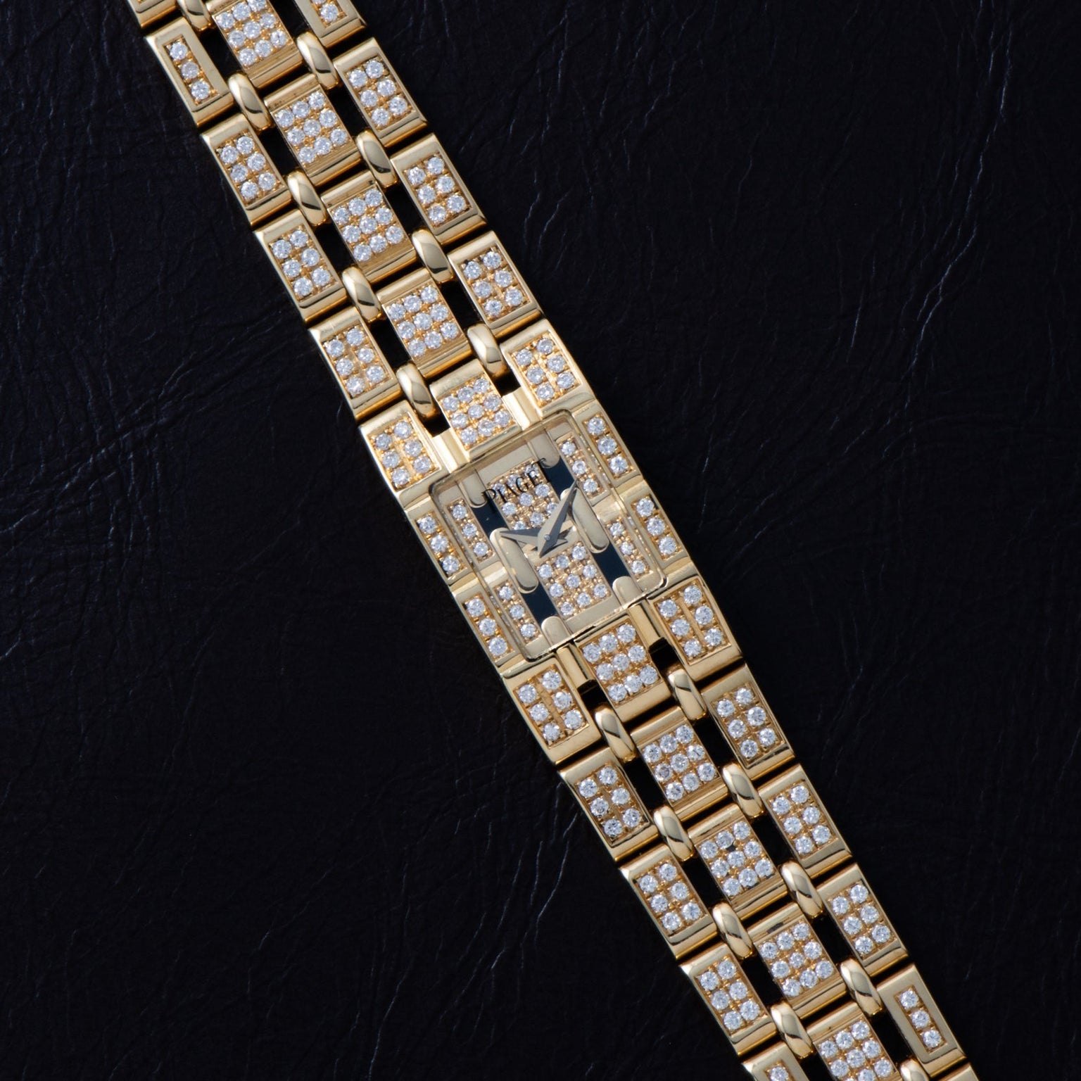 The Piaget Polo Pave, a gold wristwatch with a rectangular face and diamond accents, is showcased on a black leather surface.