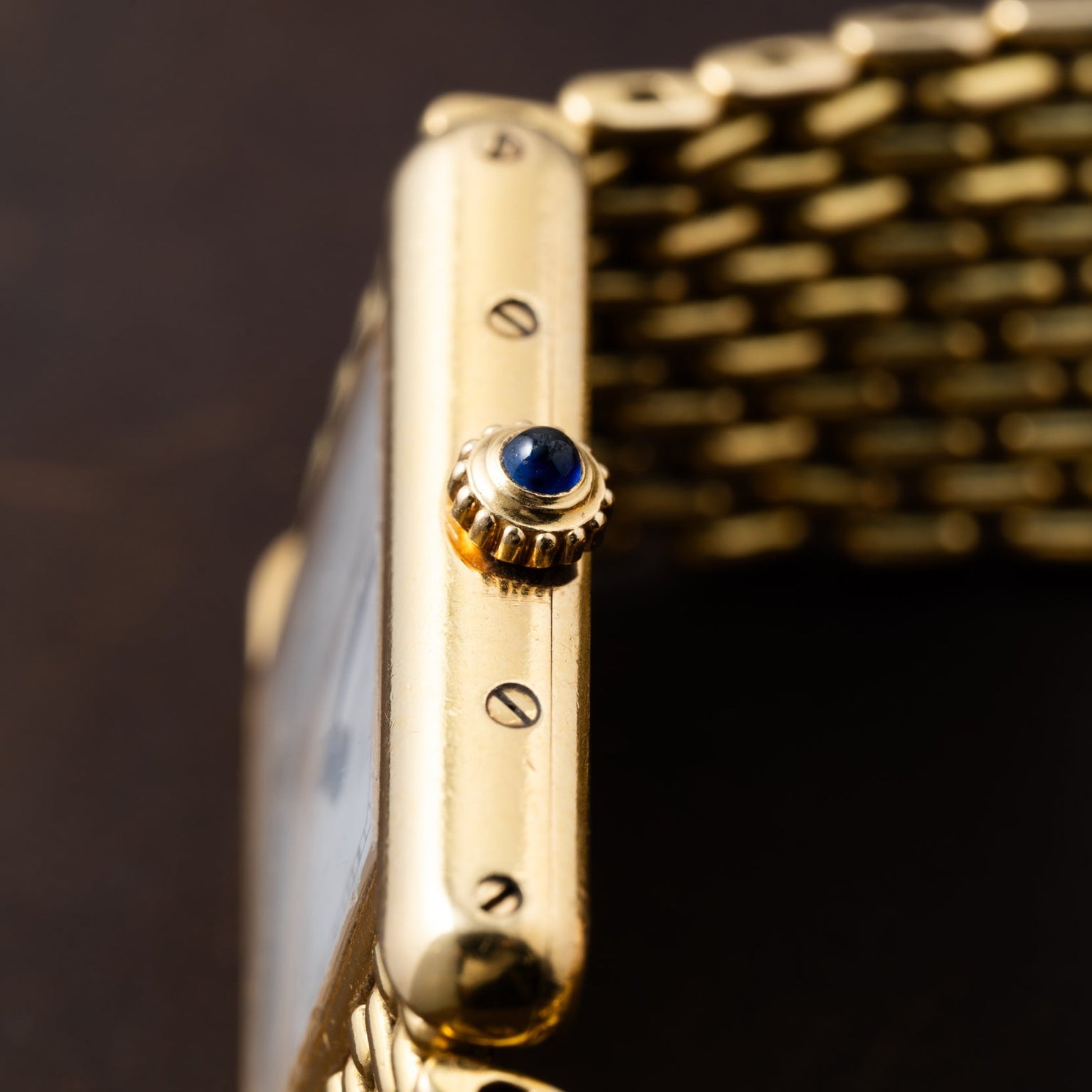 Close-up of the Cartier Tank Louis gold wristwatch crown set with a blue gemstone and its metal link bracelet.