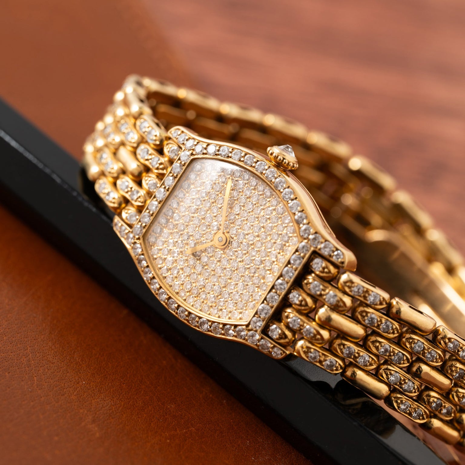 The Cartier Tortue Diamond wristwatch, adorned with diamonds, is displayed on brown leather and a black surface.