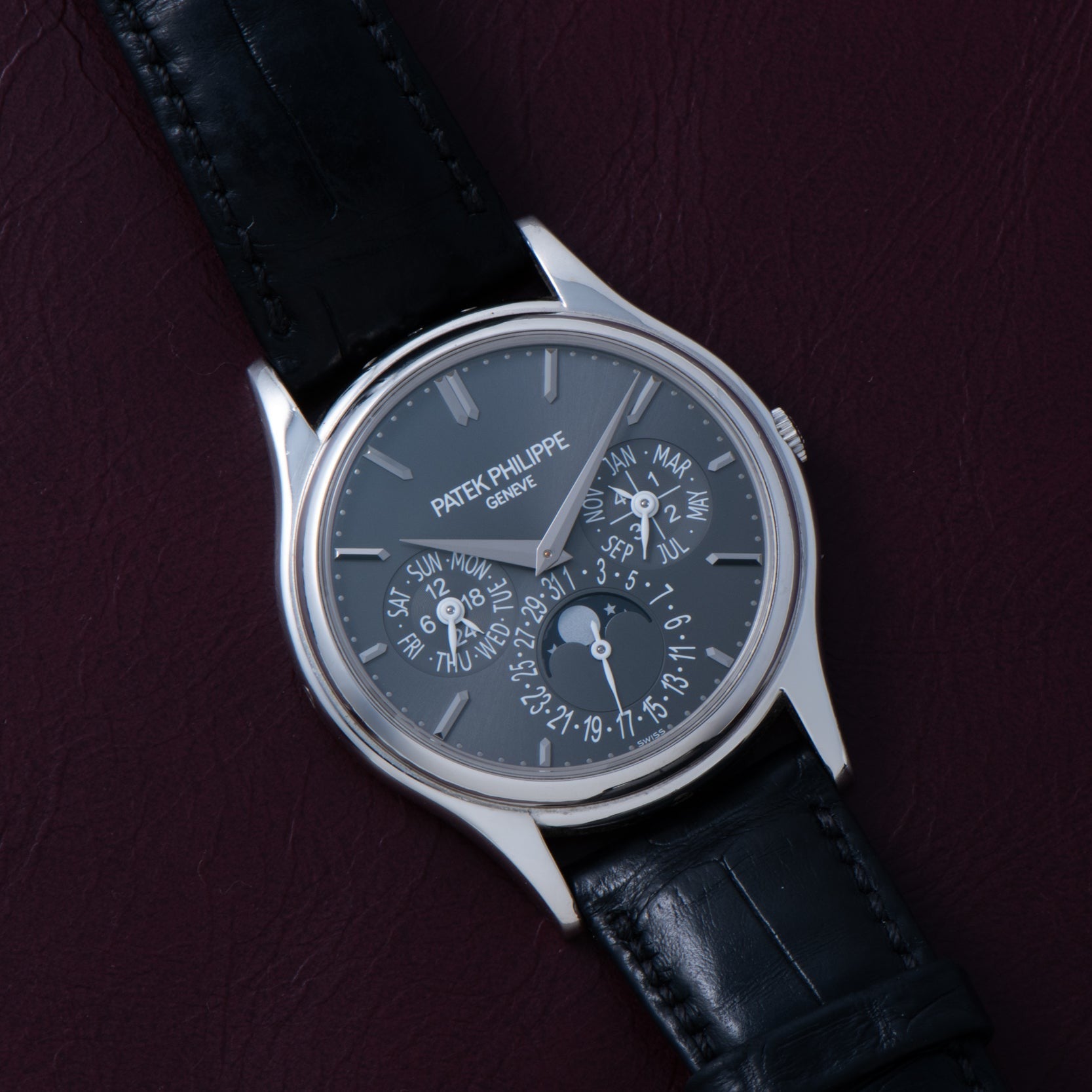 The Patek Philippe Perpetual Calendar wristwatch with a black leather strap is displayed on a dark maroon background.