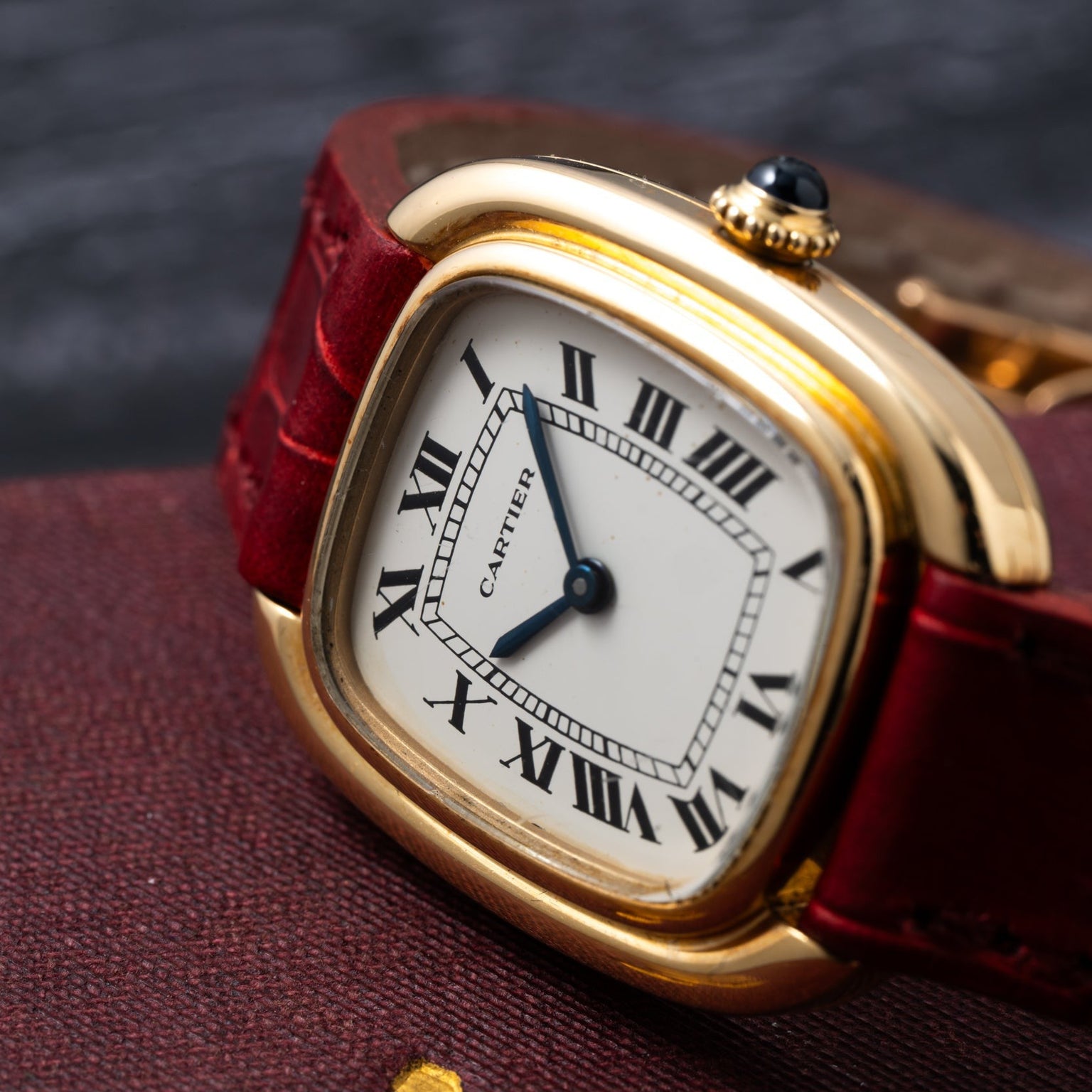 The Cartier Gondole Ladies watch features a gold case, red leather strap, and Roman numerals, displayed on a textured dark surface.