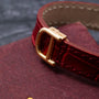 Close-up of the gold buckle on a Cartier Gondole Ladies red leather watch strap, resting on a textured maroon surface.