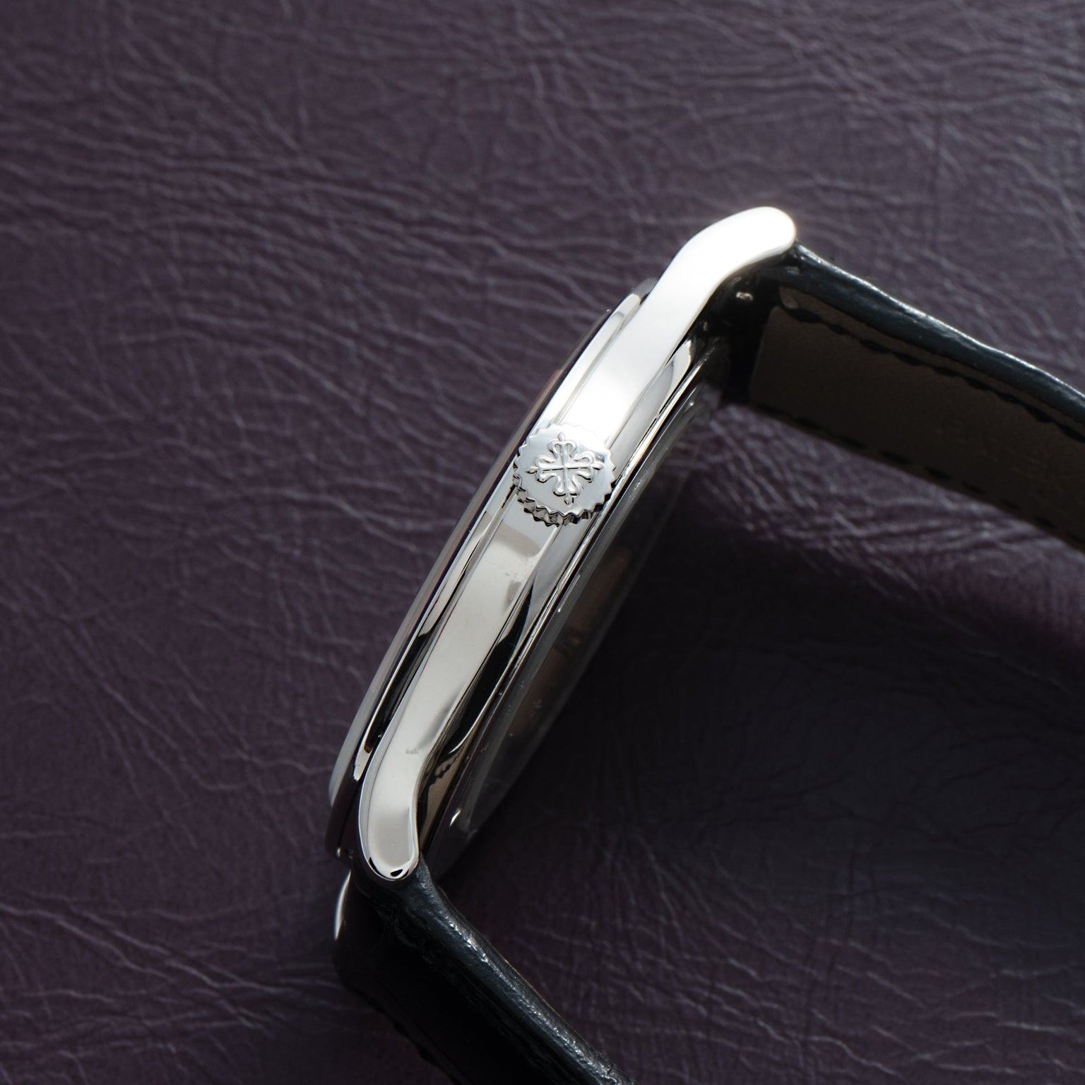 Side view of the Patek Philippe Calatrava Cloisonné Iris & Night Fireflies silver wristwatch with a black leather strap resting on a dark textured surface.