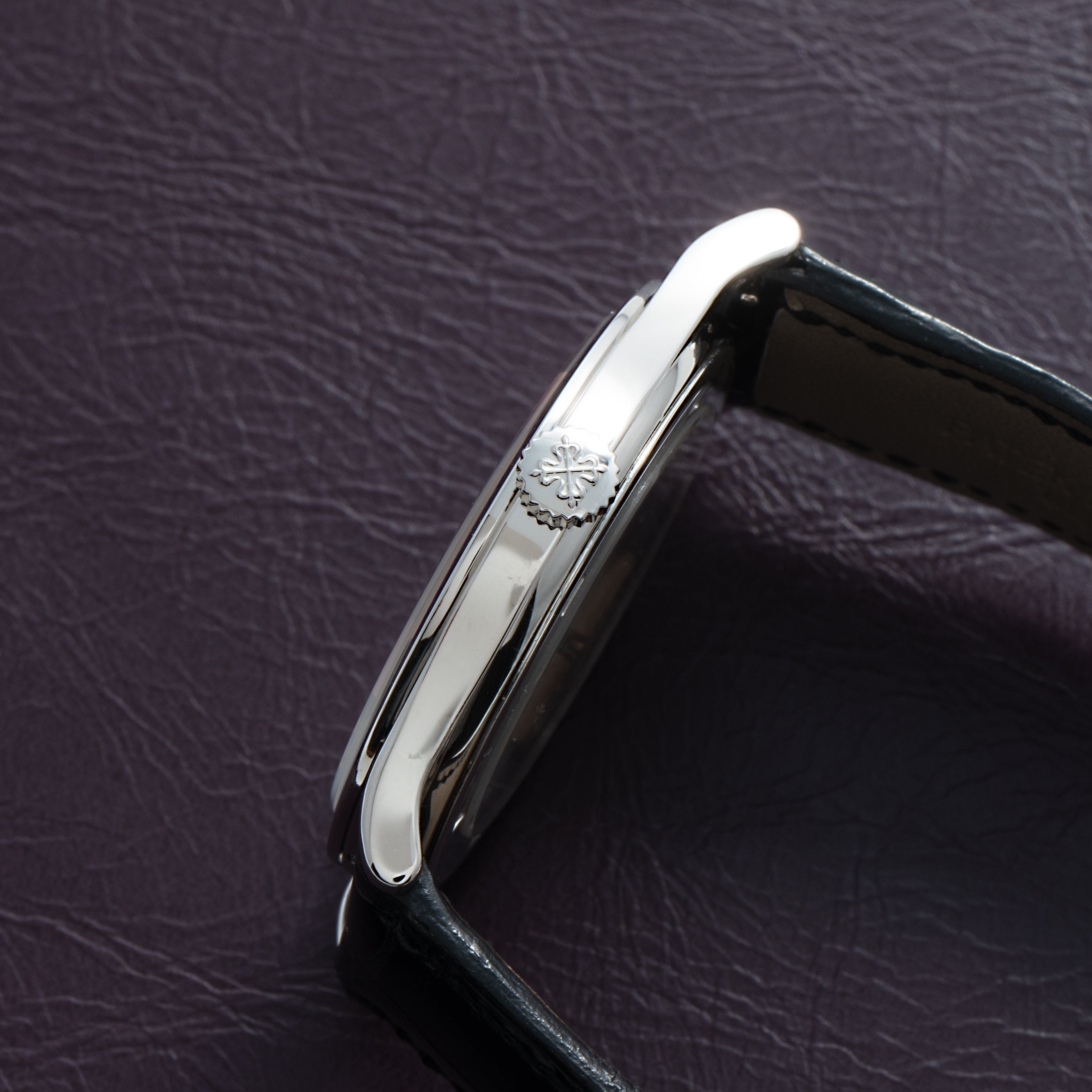 Side view of the Patek Philippe Calatrava Cloisonné Iris & Night Fireflies silver wristwatch with a black leather strap resting on a dark textured surface.