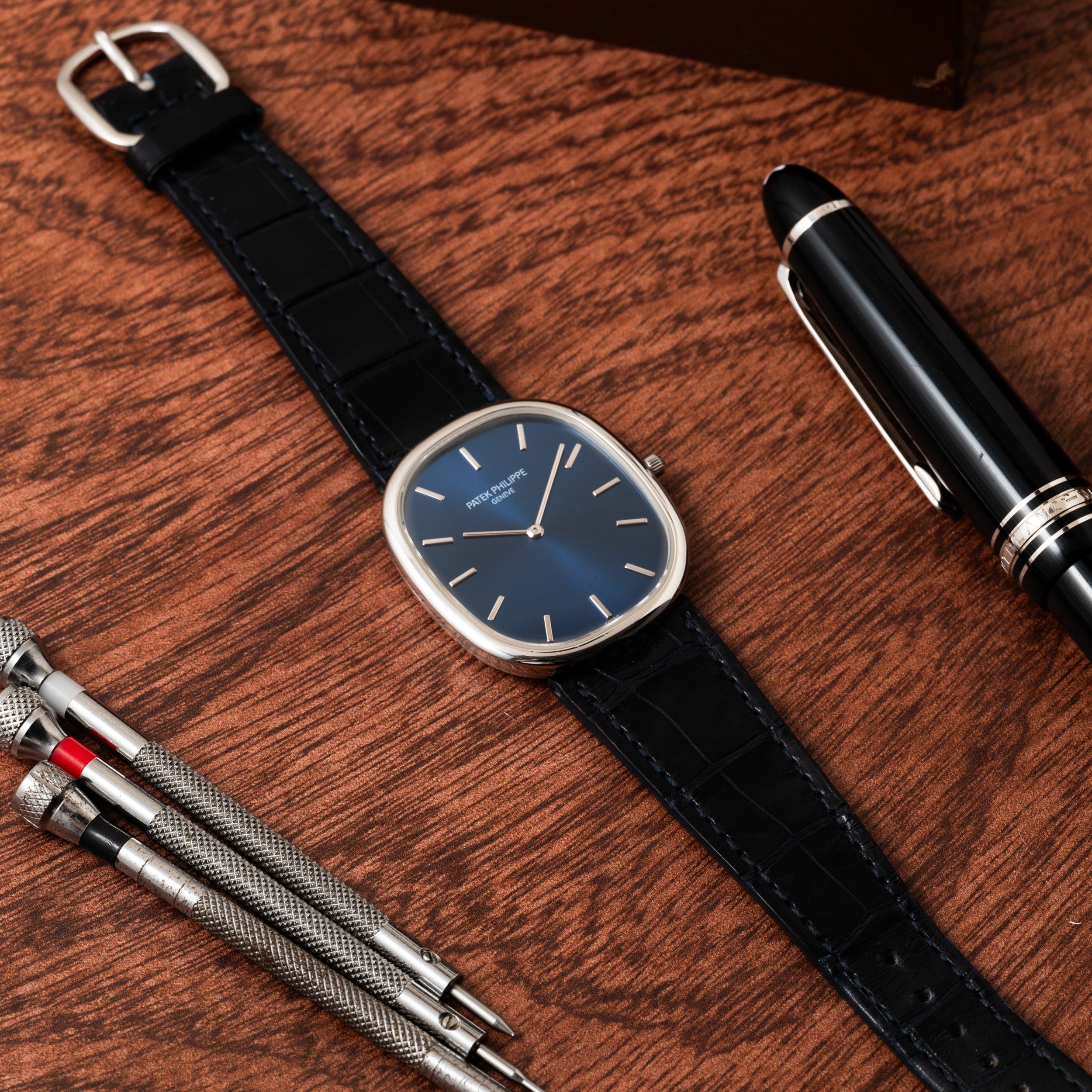 A Patek Philippe Ellipse Jumbo wristwatch, a pen, and small metal tools arranged on a wooden surface.