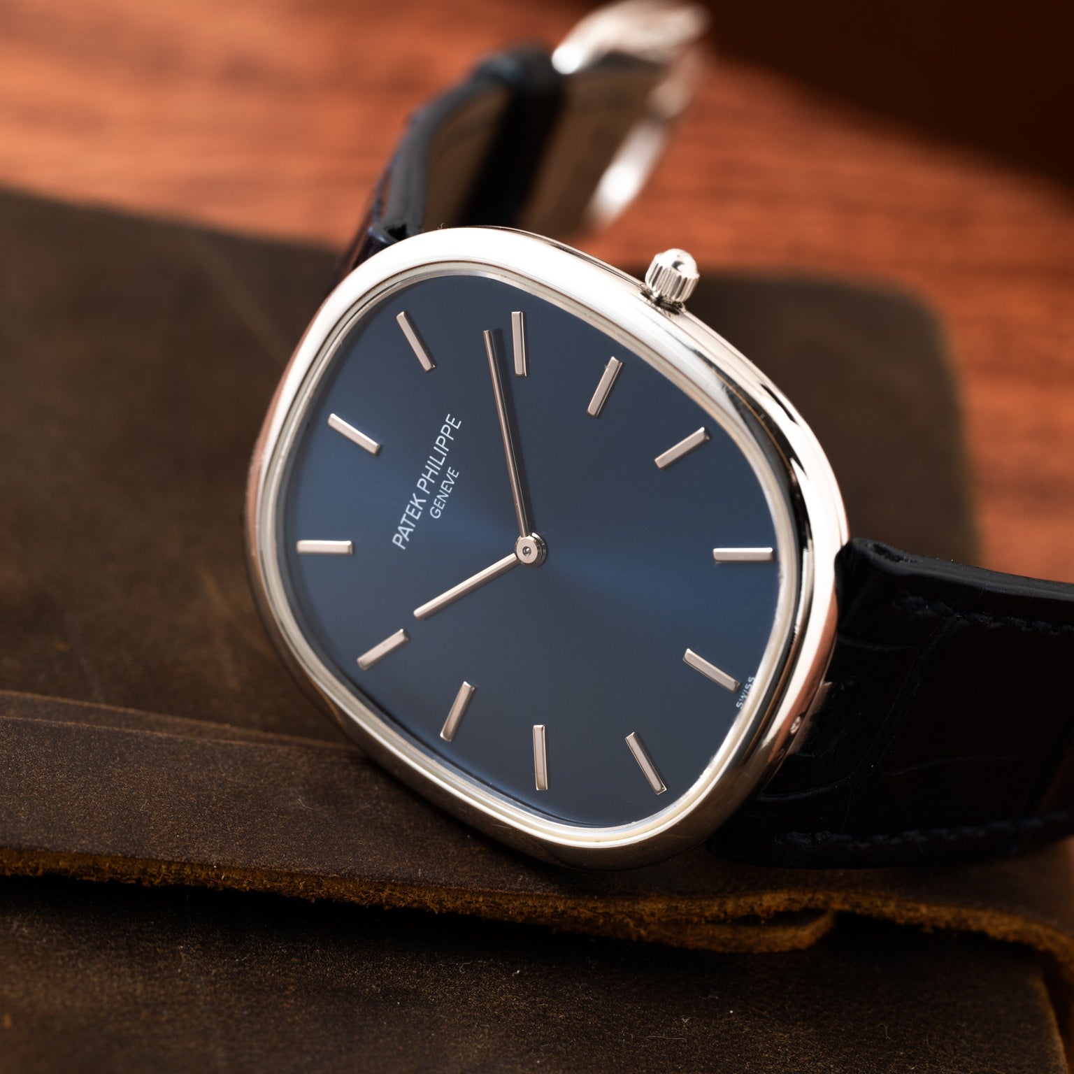 The Patek Philippe Ellipse Jumbo watch features a blue face and black leather strap, elegantly displayed on a brown surface.
