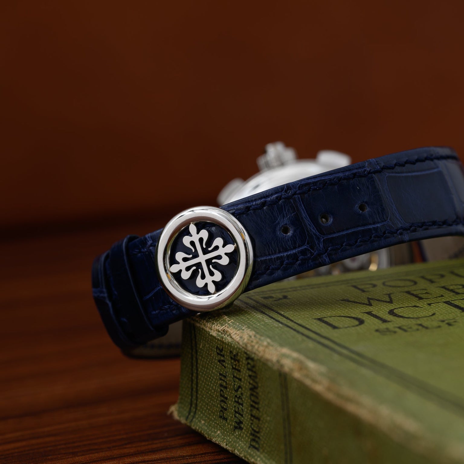 The Patek Philippe Split-Seconds Chronograph, a dark blue luxury watch with a silver emblem, rests on a green hardcover book against a brown background.