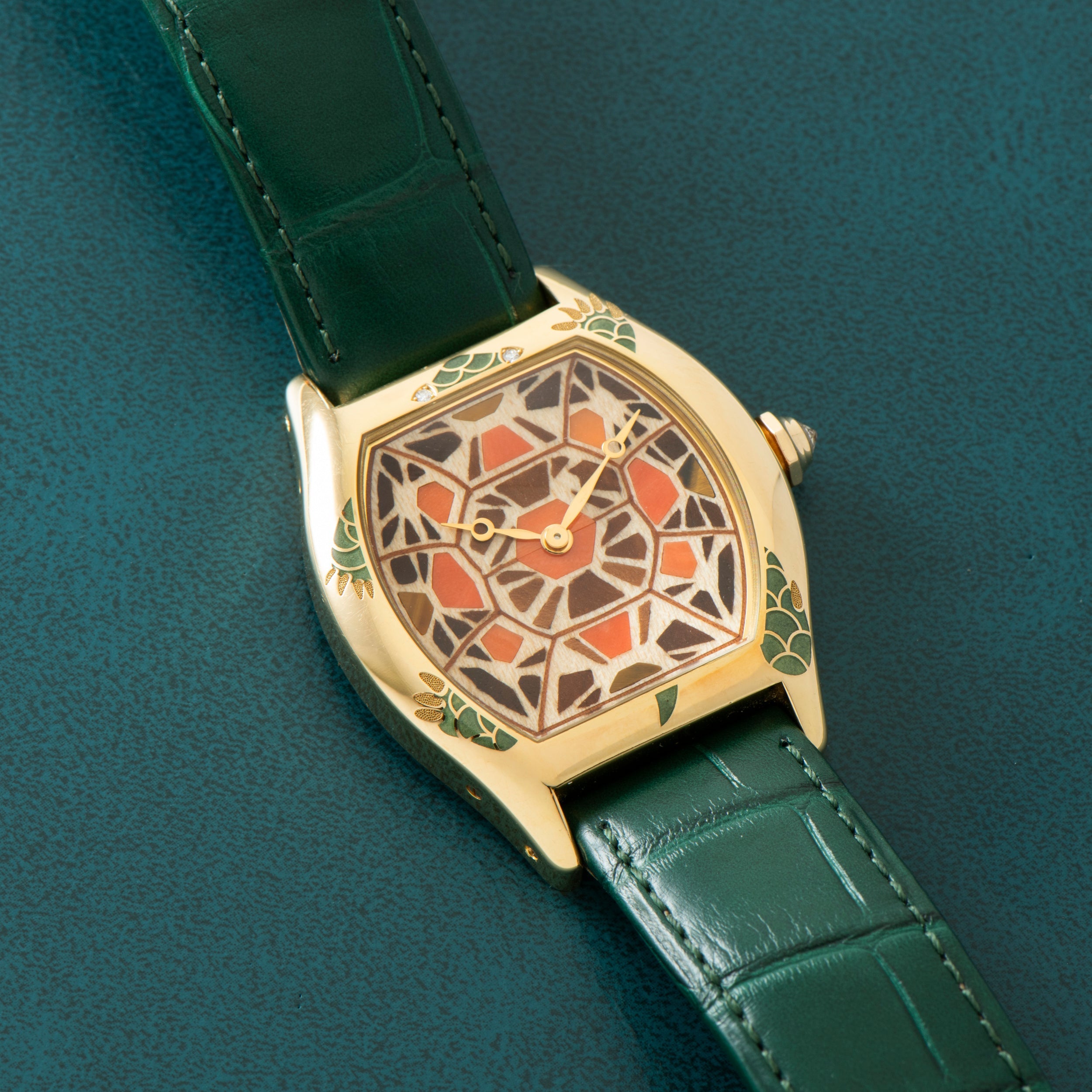 The Cartier Tortue XL Turtle Limited Edition gold watch features a green leather strap and a geometric mosaic-patterned face, shown on a green surface.