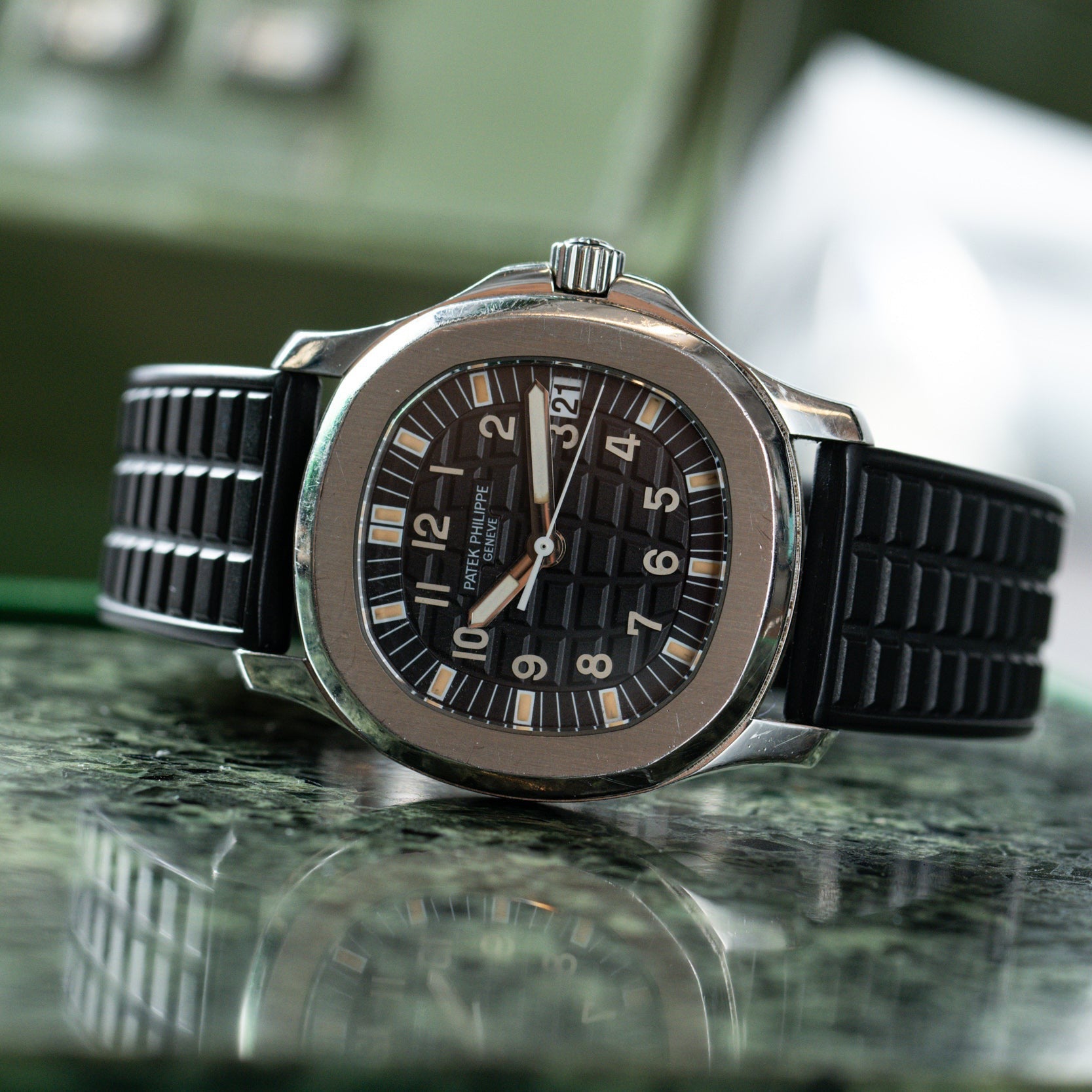 The Patek Philippe Aquanaut, a silver wristwatch with a black rubber strap, is displayed on a green marble surface.