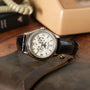 A Patek Philippe Annual Calendar Moonphase wristwatch with a black leather strap rests on a brown case, while an old beige telephone and a book are blurred in the background. The watch features an annual calendar, day, date, and 24-hour indicators.