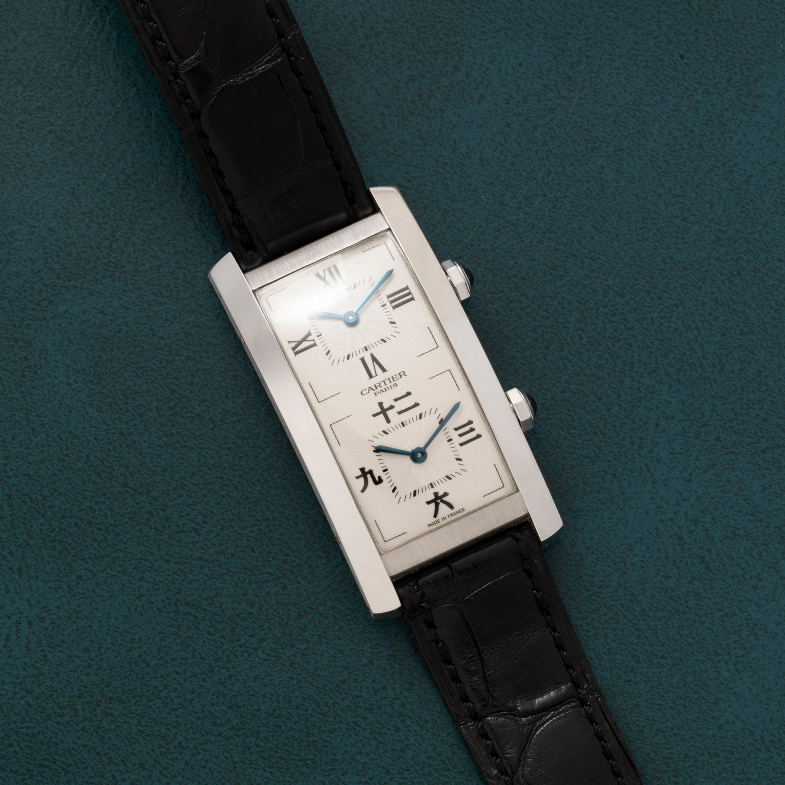 A Cartier Tank Cintrée CPCP Dual-Time wristwatch with a rectangular case, two dials, and a black leather strap on a green textured surface.